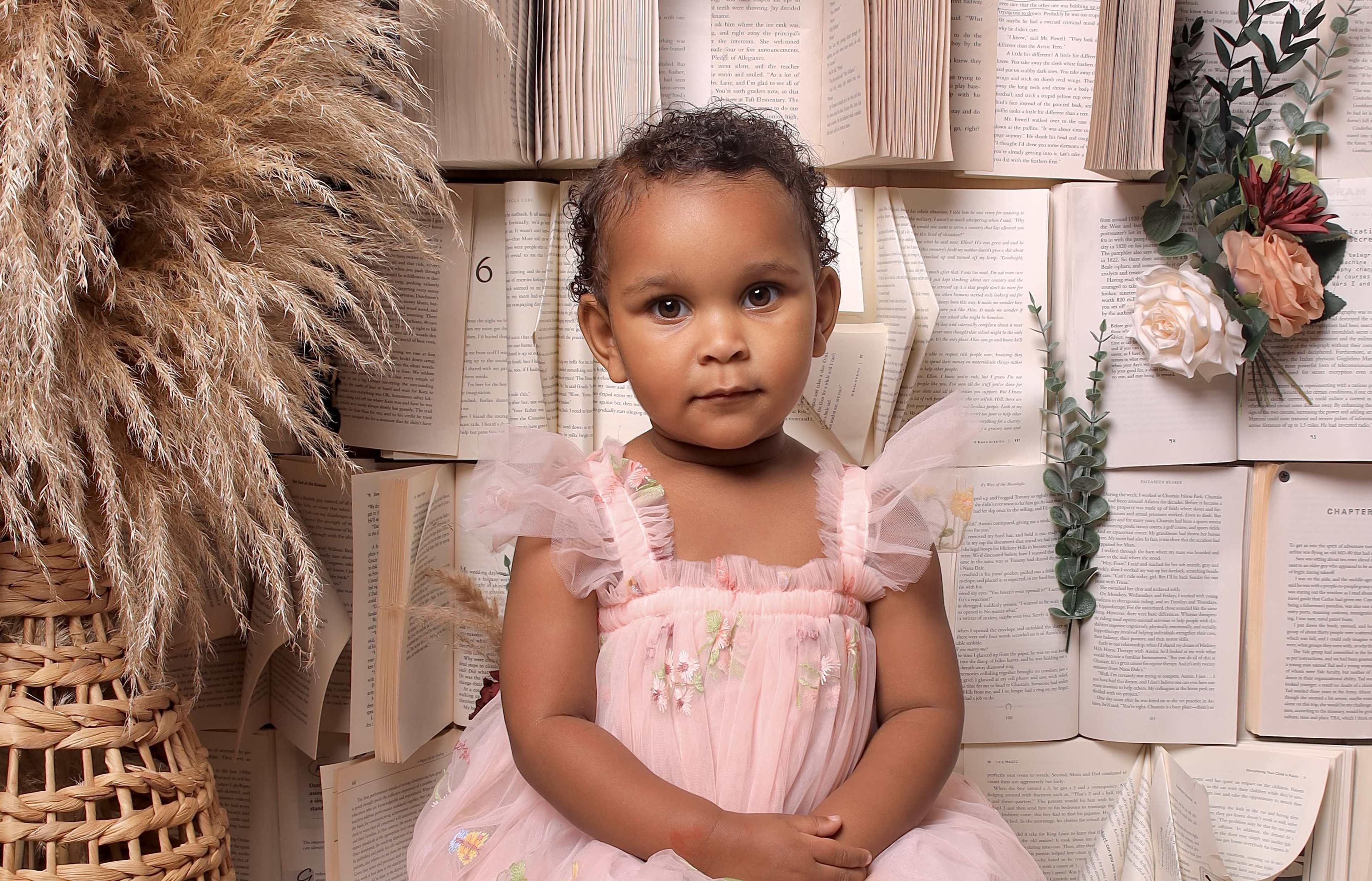 A young child wearing a pink dress sits in front of a backdrop made of open books surrounded by decorative plants.