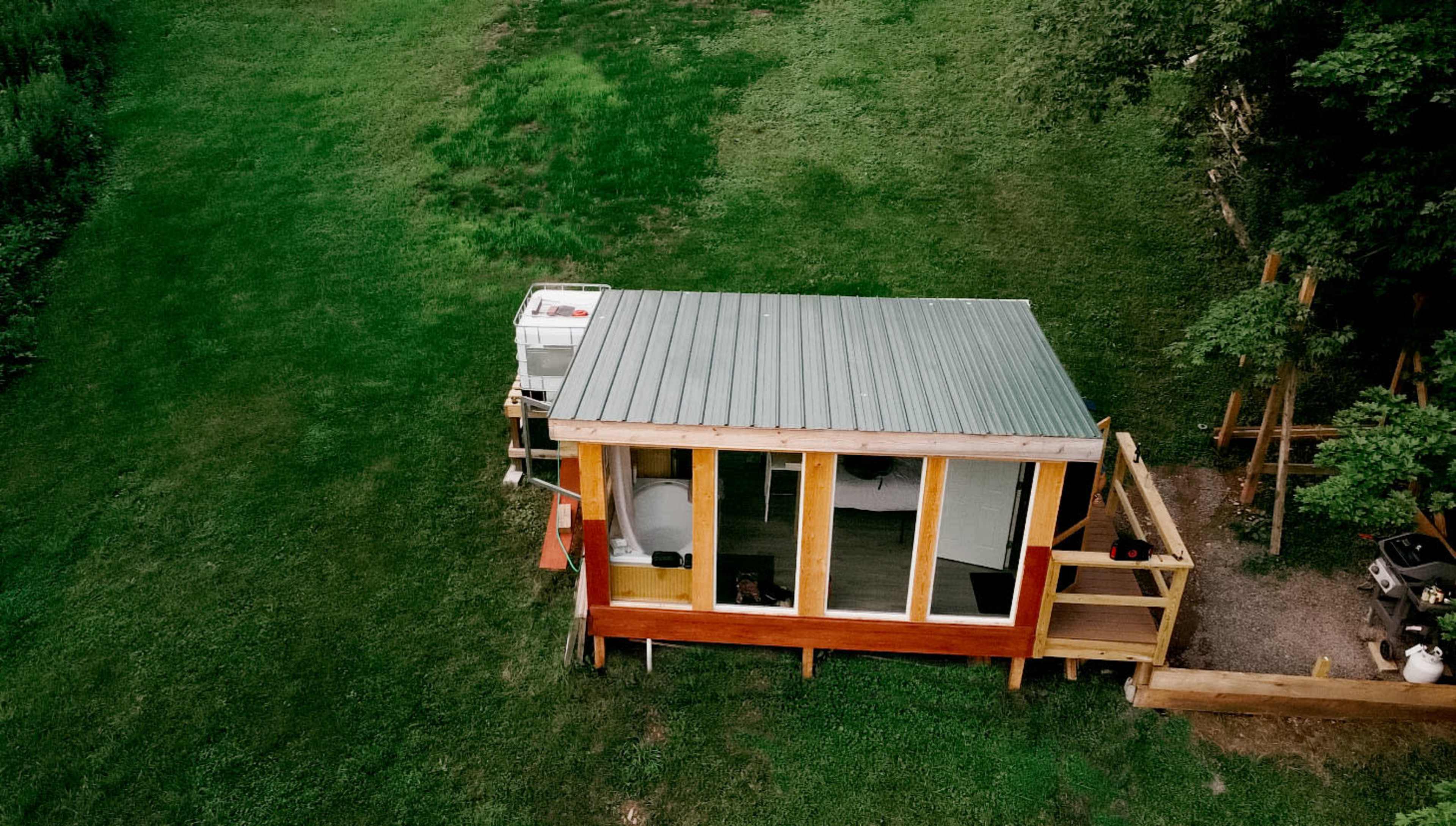 A small wooden structure with large glass windows and a metal roof, situated in a grassy area.