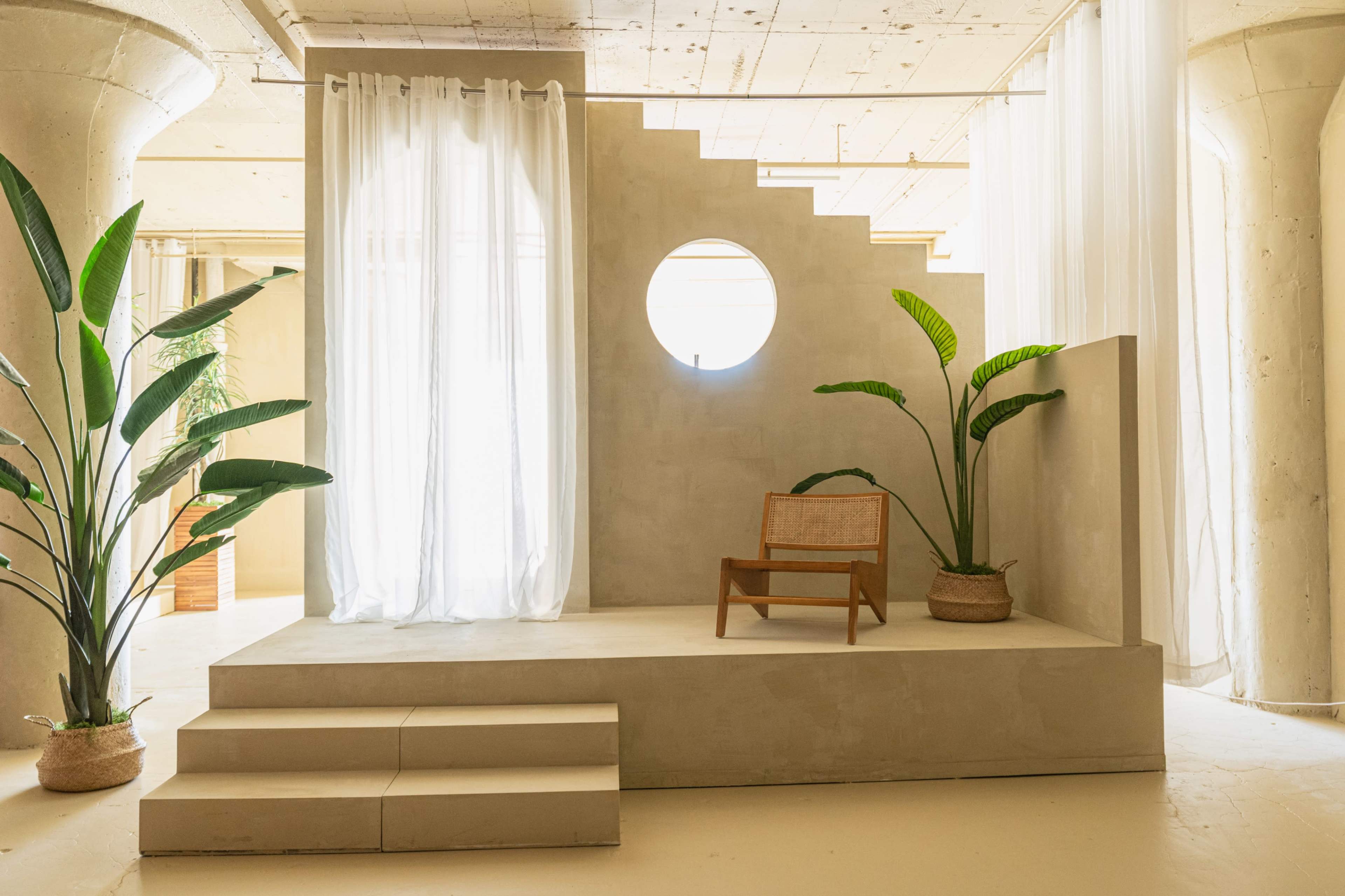 A minimalist interior space featuring a stepped platform, a circular window, sheer curtains, and two potted plants alongside a simple wooden chair.