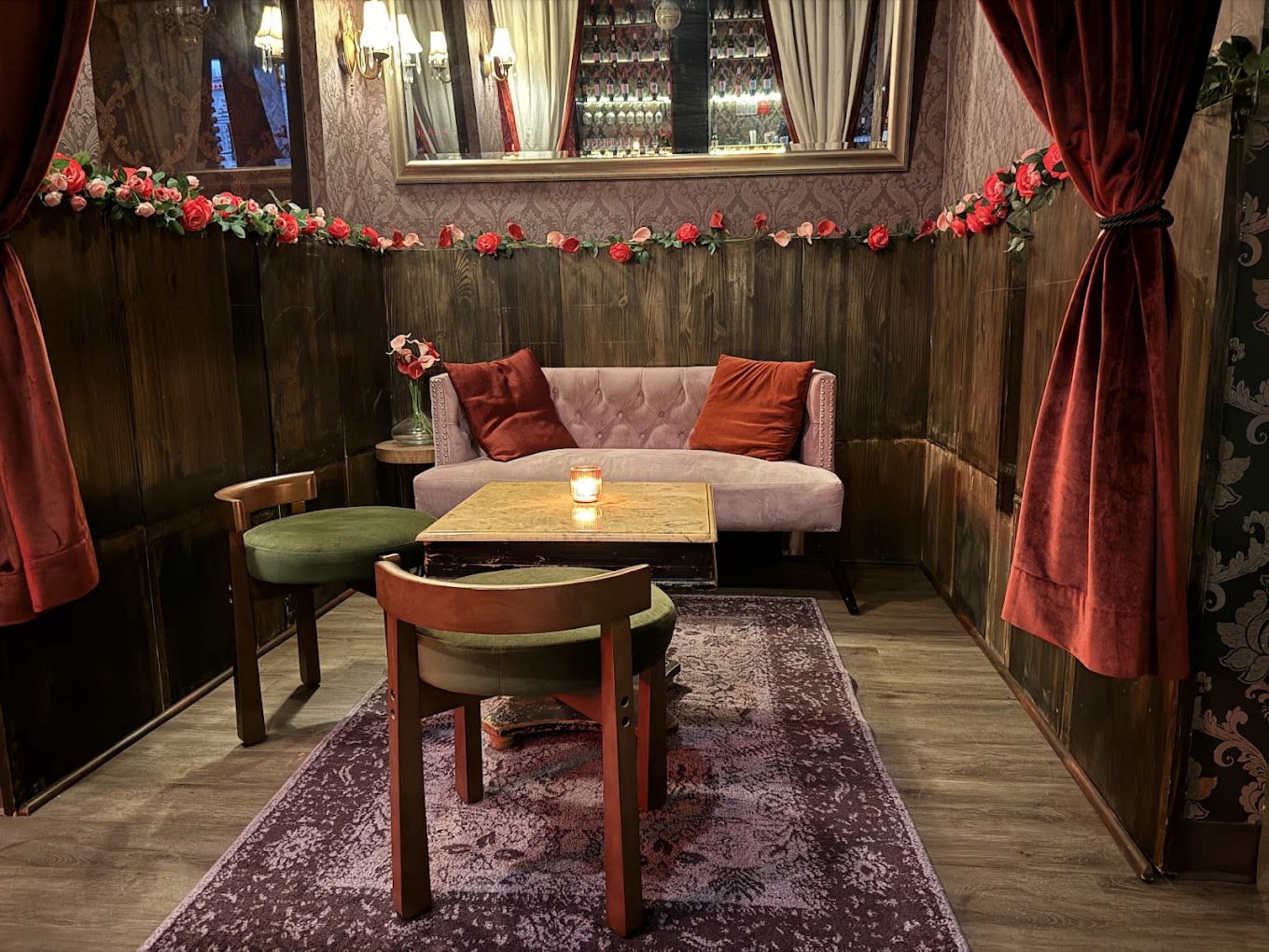 The image shows a cozy seating area with a pink sofa, a small wooden table, and two chairs, all surrounded by decorative flowers and warm lighting.