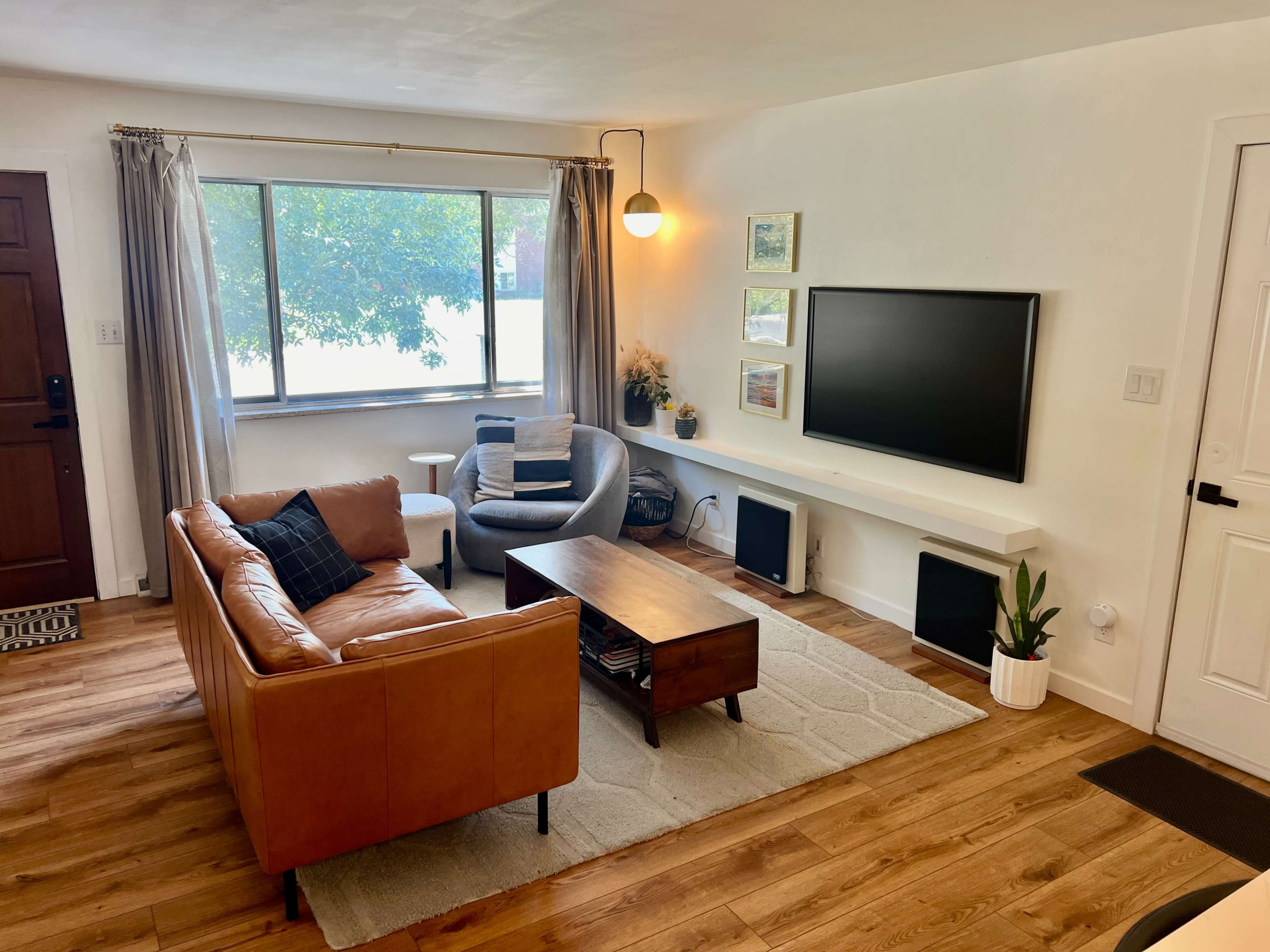 The living room features a tan leather sofa, a wooden coffee table, and a large window with light curtains, framed by a TV on the wall opposite.