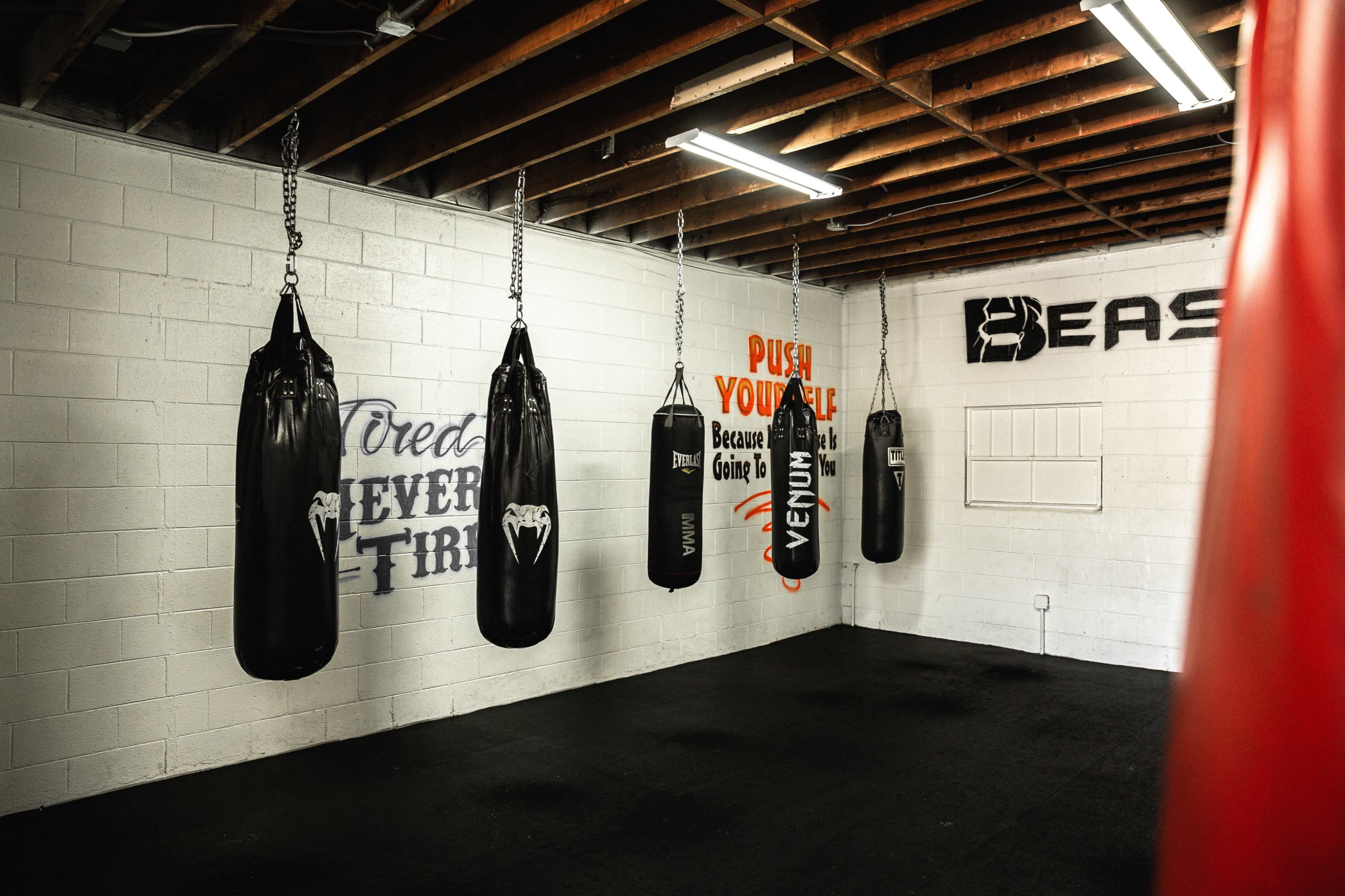 A boxing gym with several hanging punching bags, motivational wall graphics, and a black rubber floor.