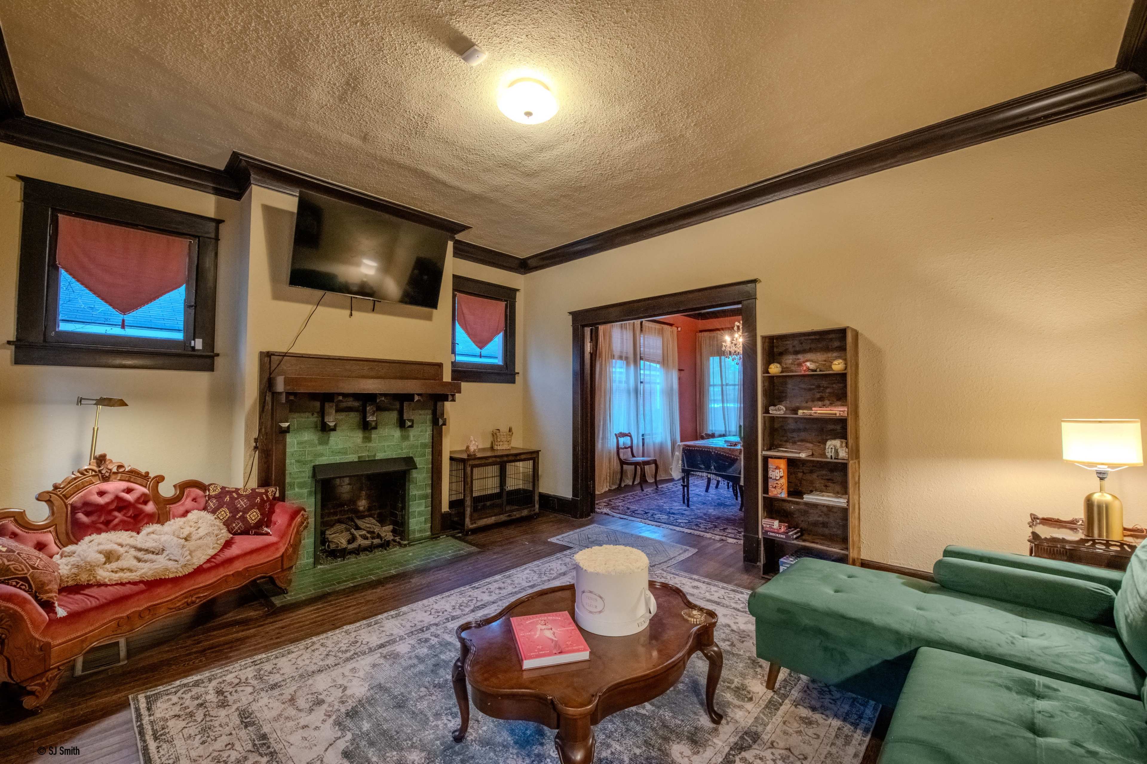 A cozy living room featuring a green fireplace, a red vintage couch, a green sofa, a television mounted on the wall, and a bookshelf filled with books.