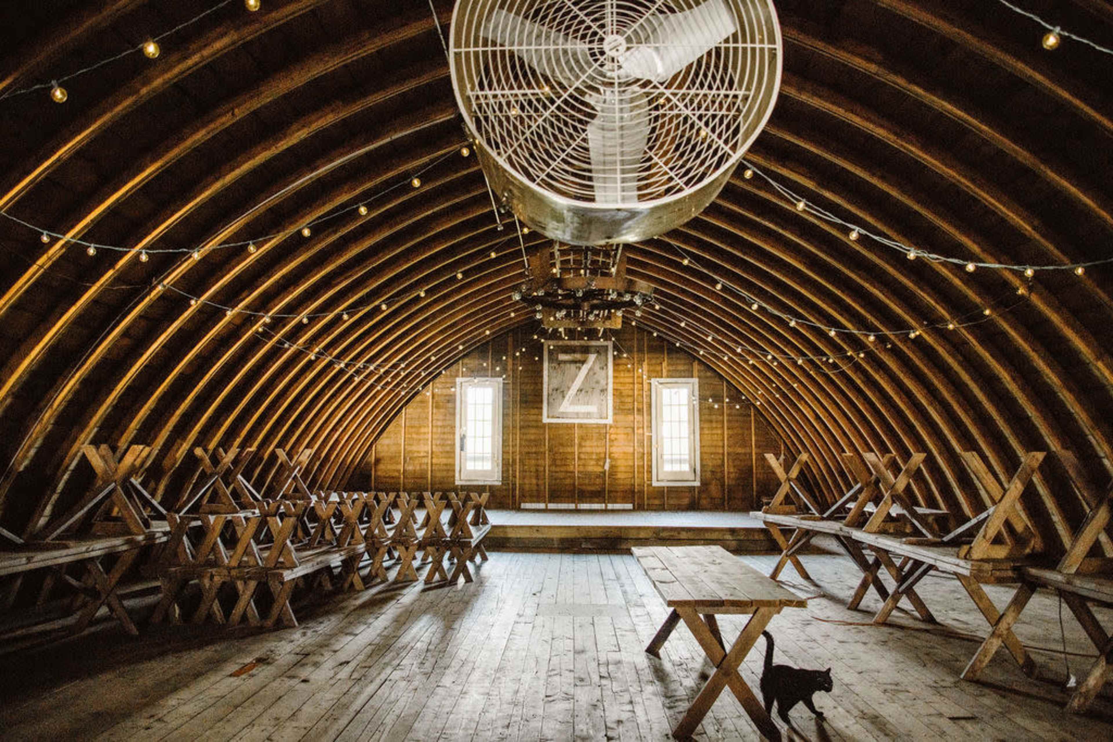 The interior of a wooden barn features arched beams, a large ceiling fan, and wooden tables arranged along the sides.