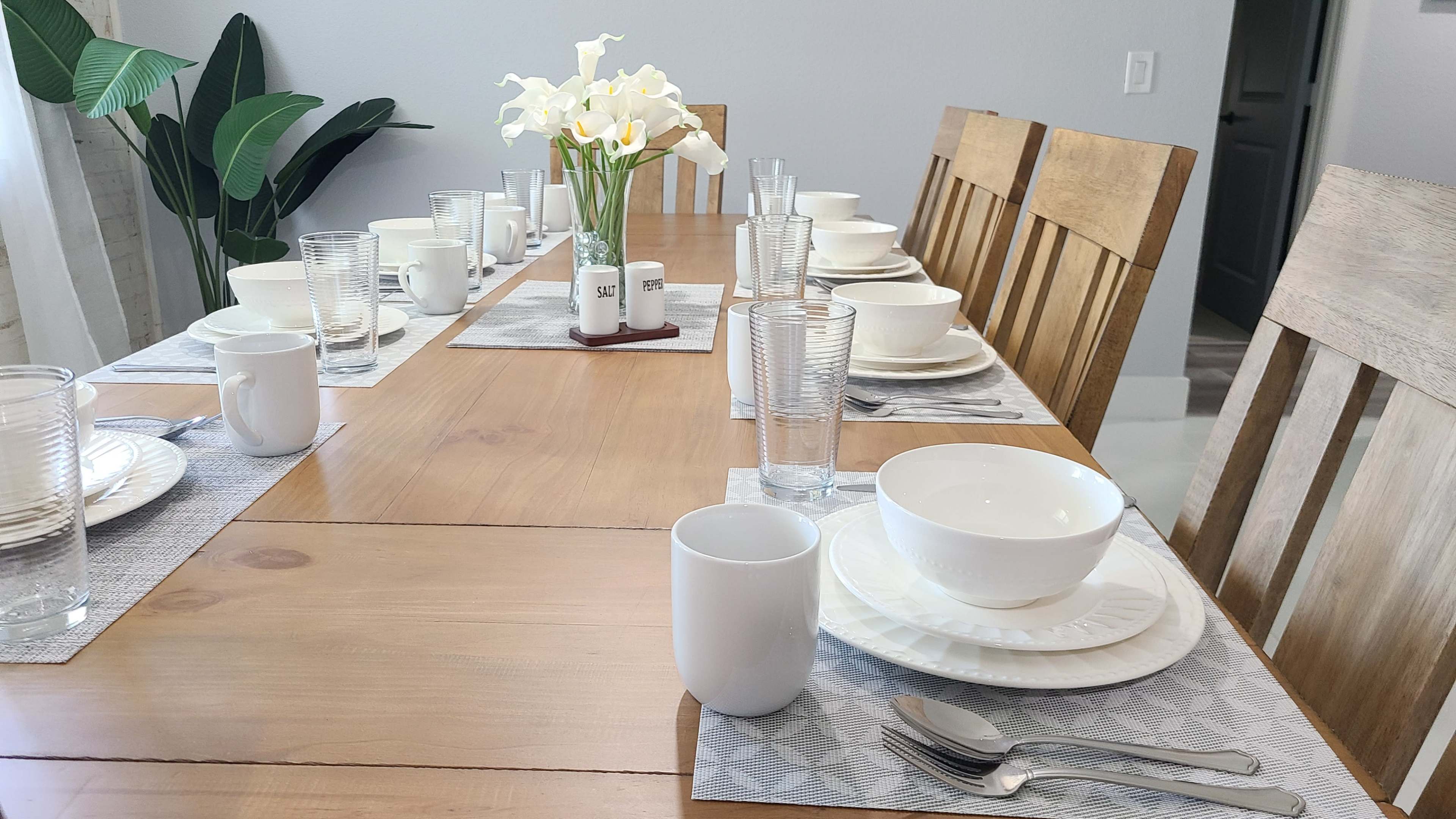 A dining table is set for a meal with plates, bowls, glasses, and a vase of flowers arranged down the center.
