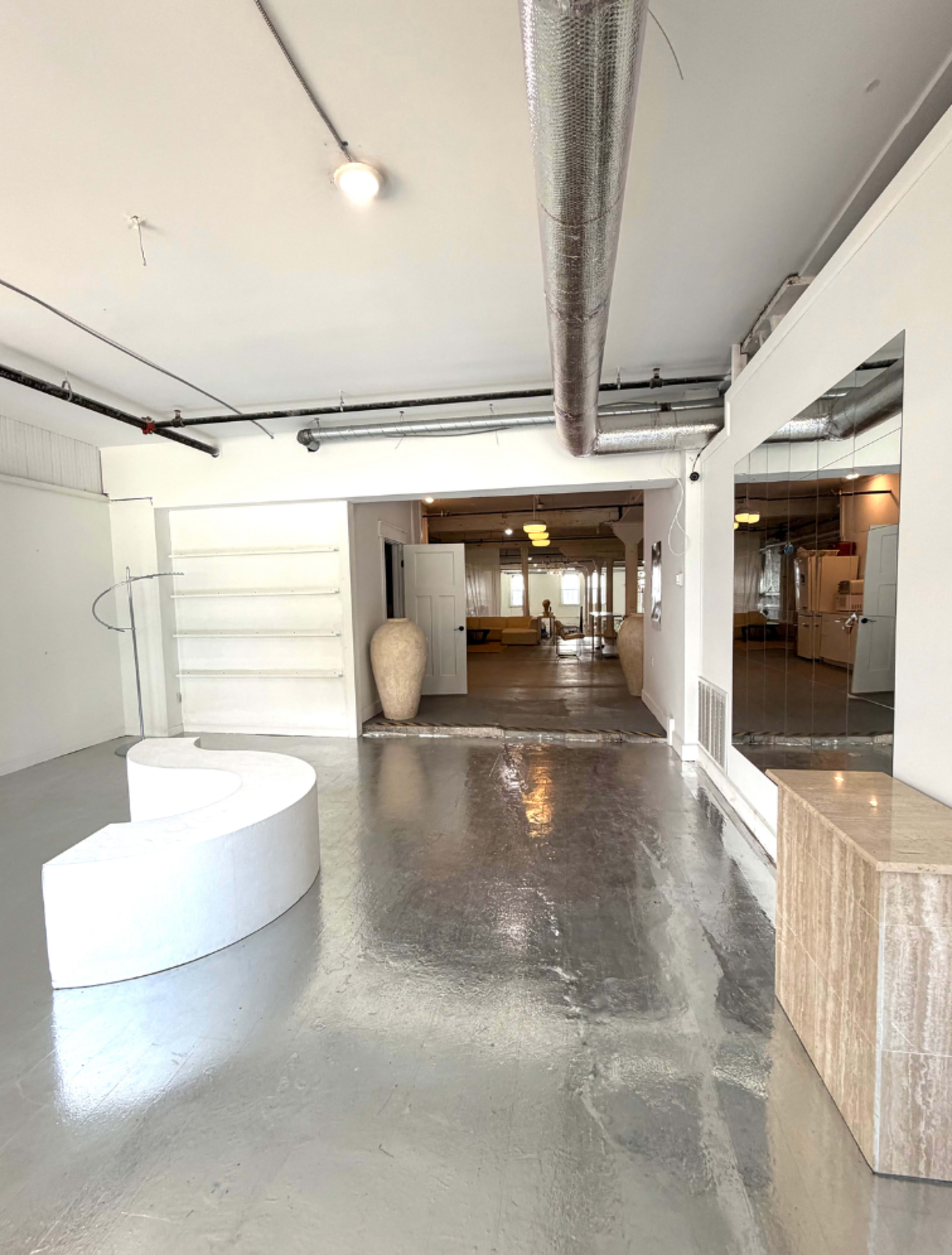 The image shows a spacious, minimalist interior with polished concrete flooring, a curved white seating area, and large mirrors reflecting the open layout.