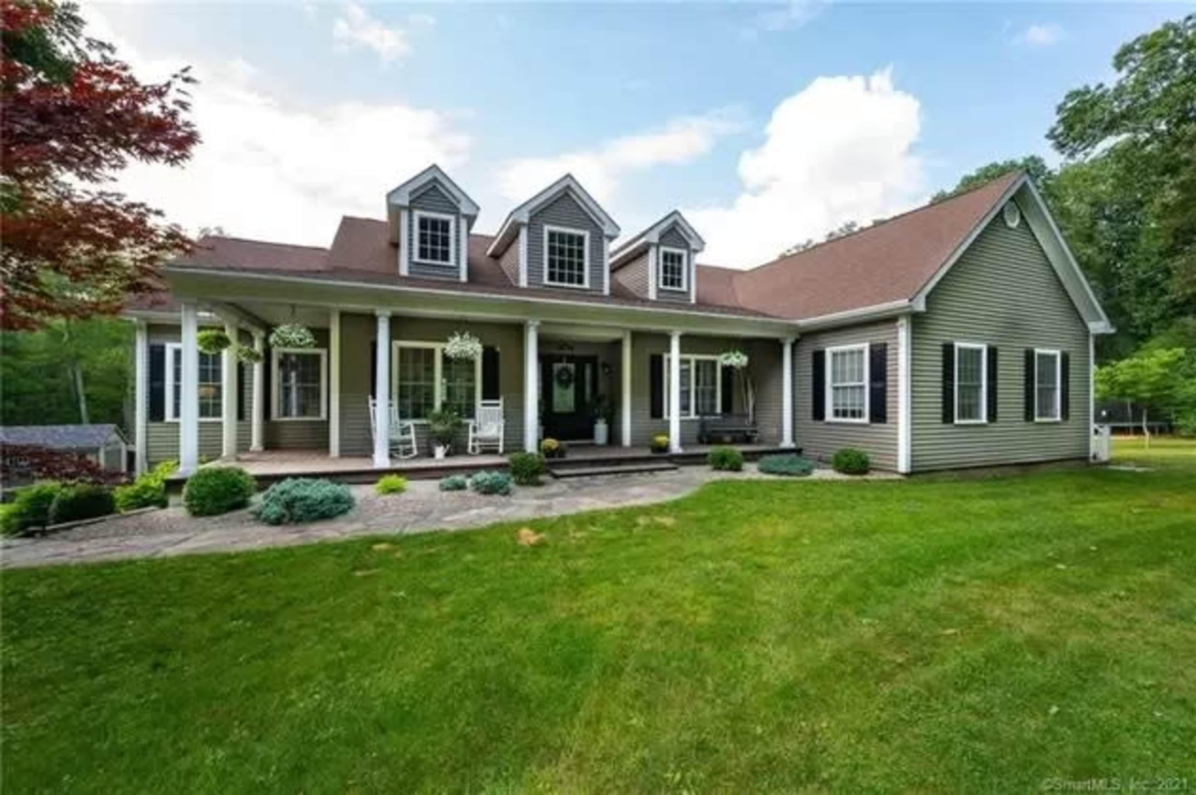 A large, two-story house with a sloped roof, featuring a front porch supported by white columns and surrounded by a green lawn and landscaping.