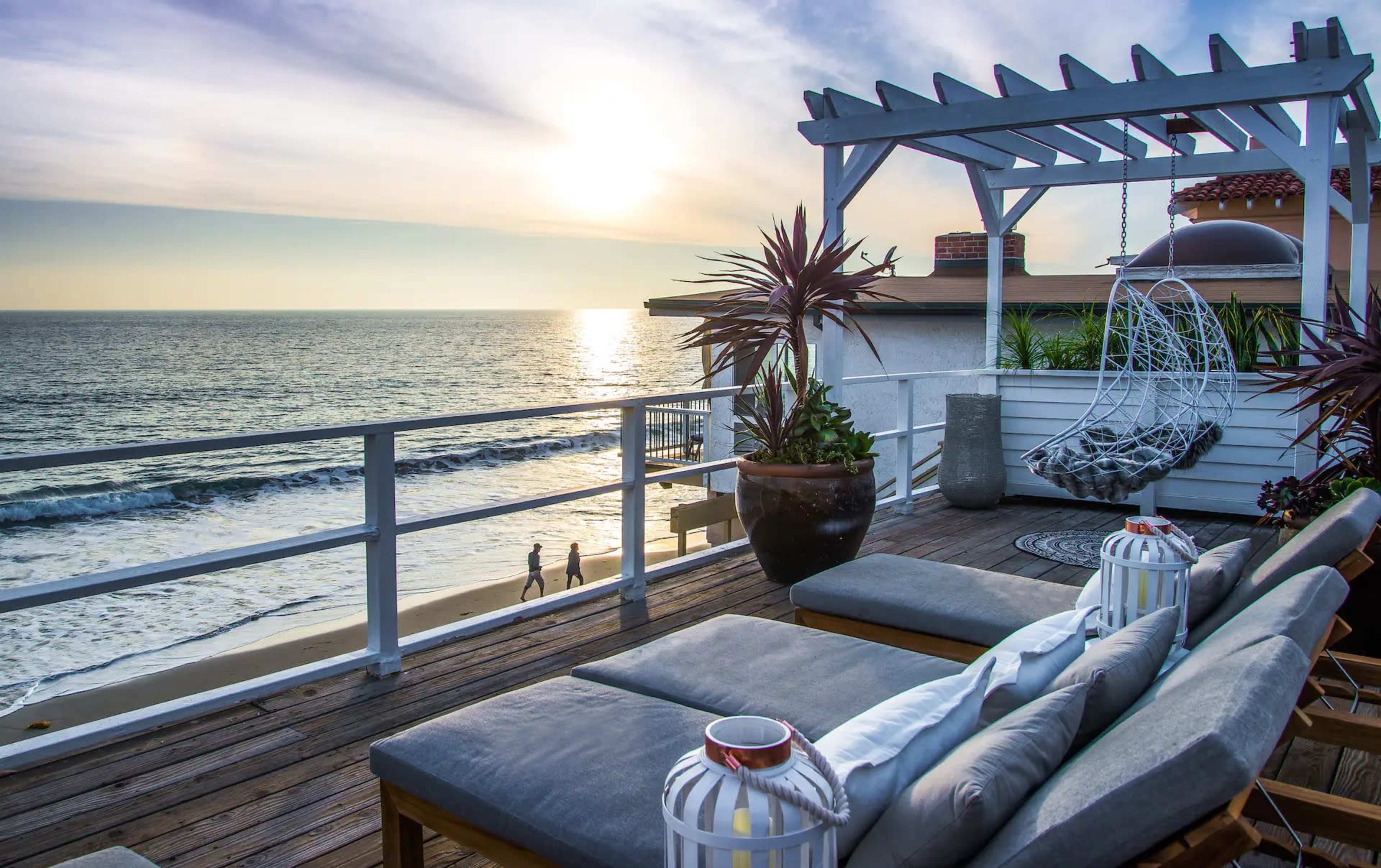 A wooden deck with lounge chairs overlooks the ocean at sunset, featuring a hanging chair and potted plants.