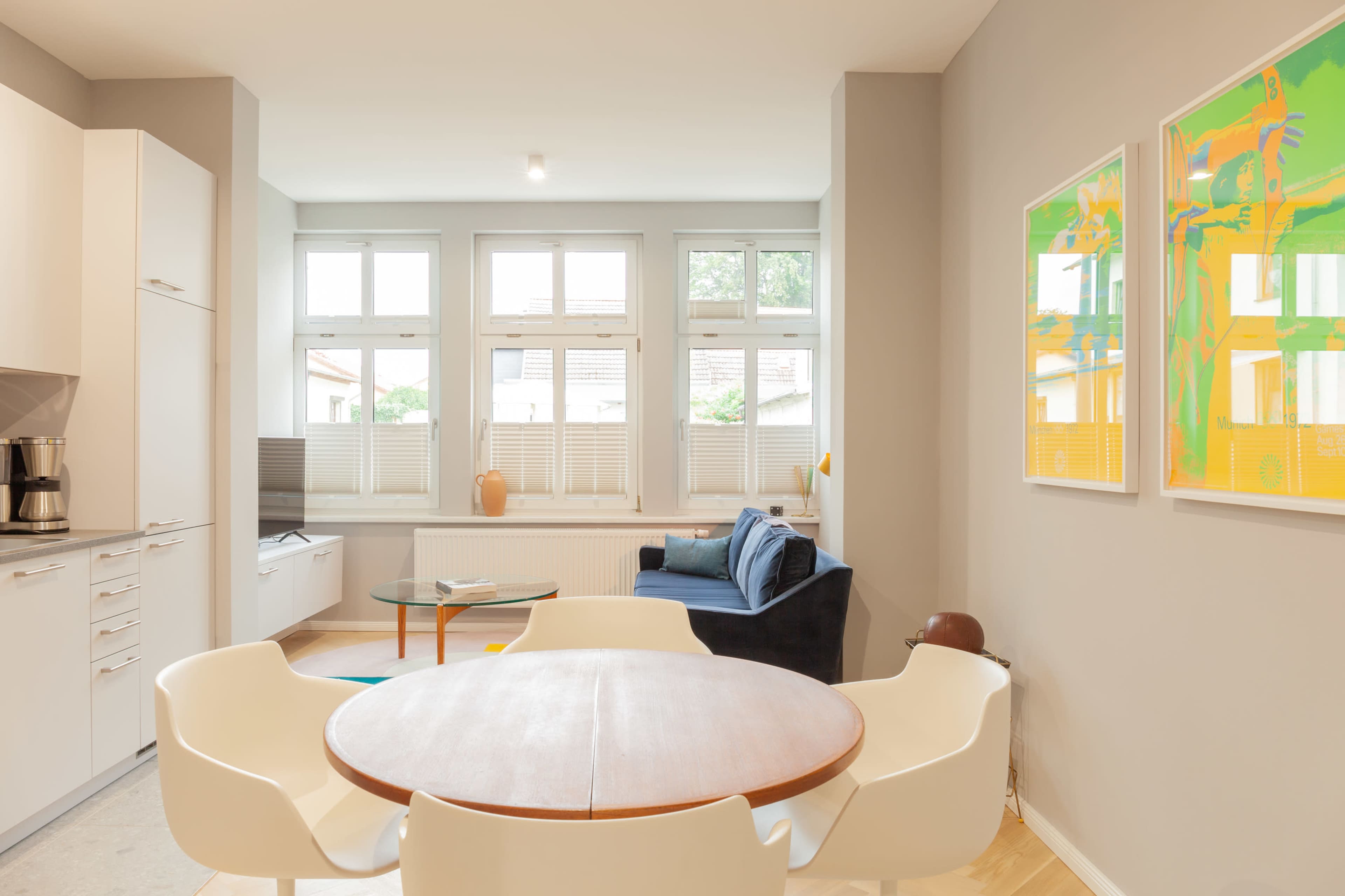 A modern living space features a kitchen area, a dining table with chairs, a sofa, and large windows allowing natural light to enter.