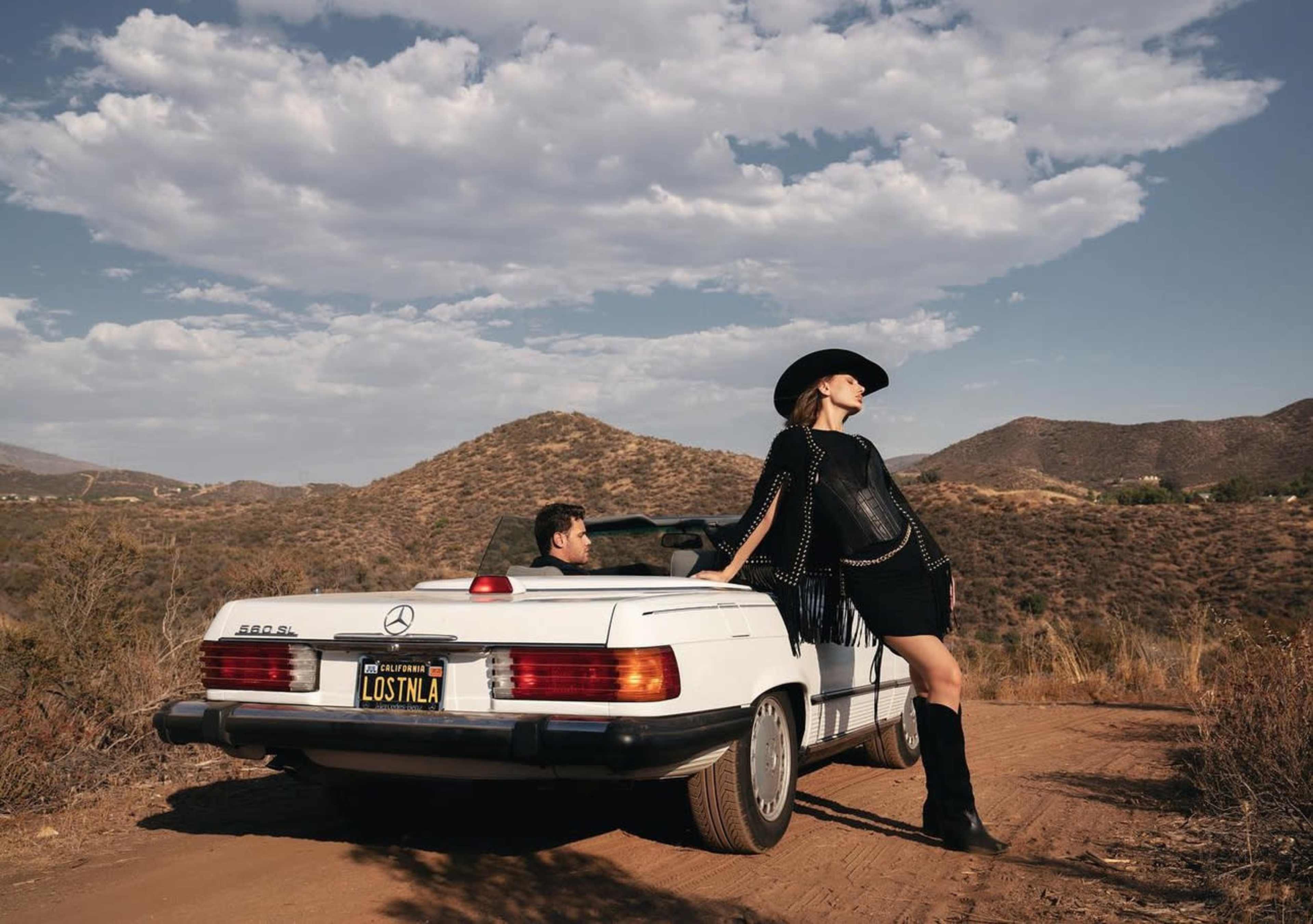 A woman in a black outfit and cowboy hat leans on a white Mercedes-Benz convertible parked on a dirt road with hills in the background.