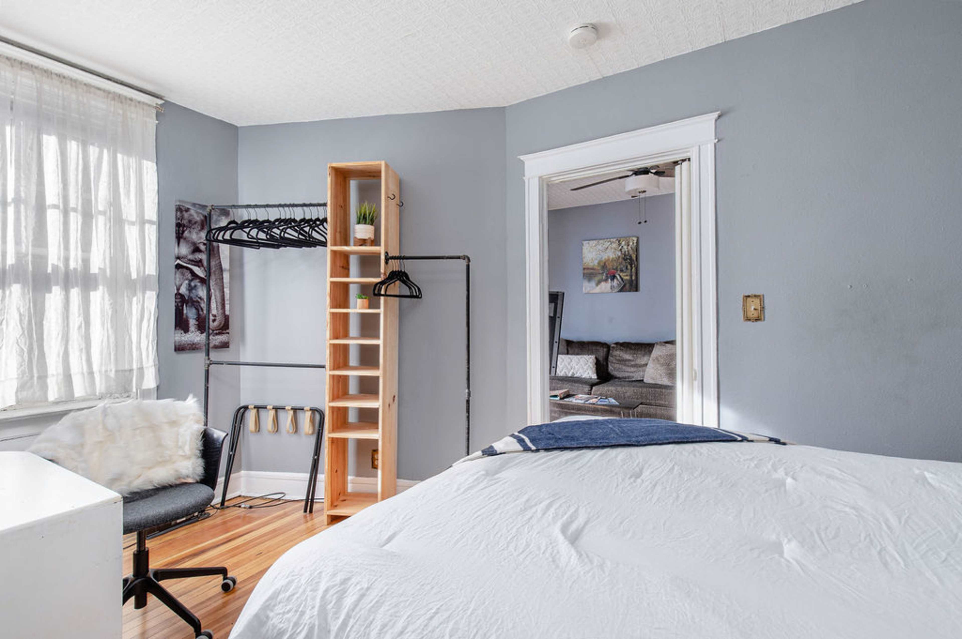 A small bedroom featuring a white bed, a desk with a chair, a clothing rack, and a shelf, with natural light coming through a window.