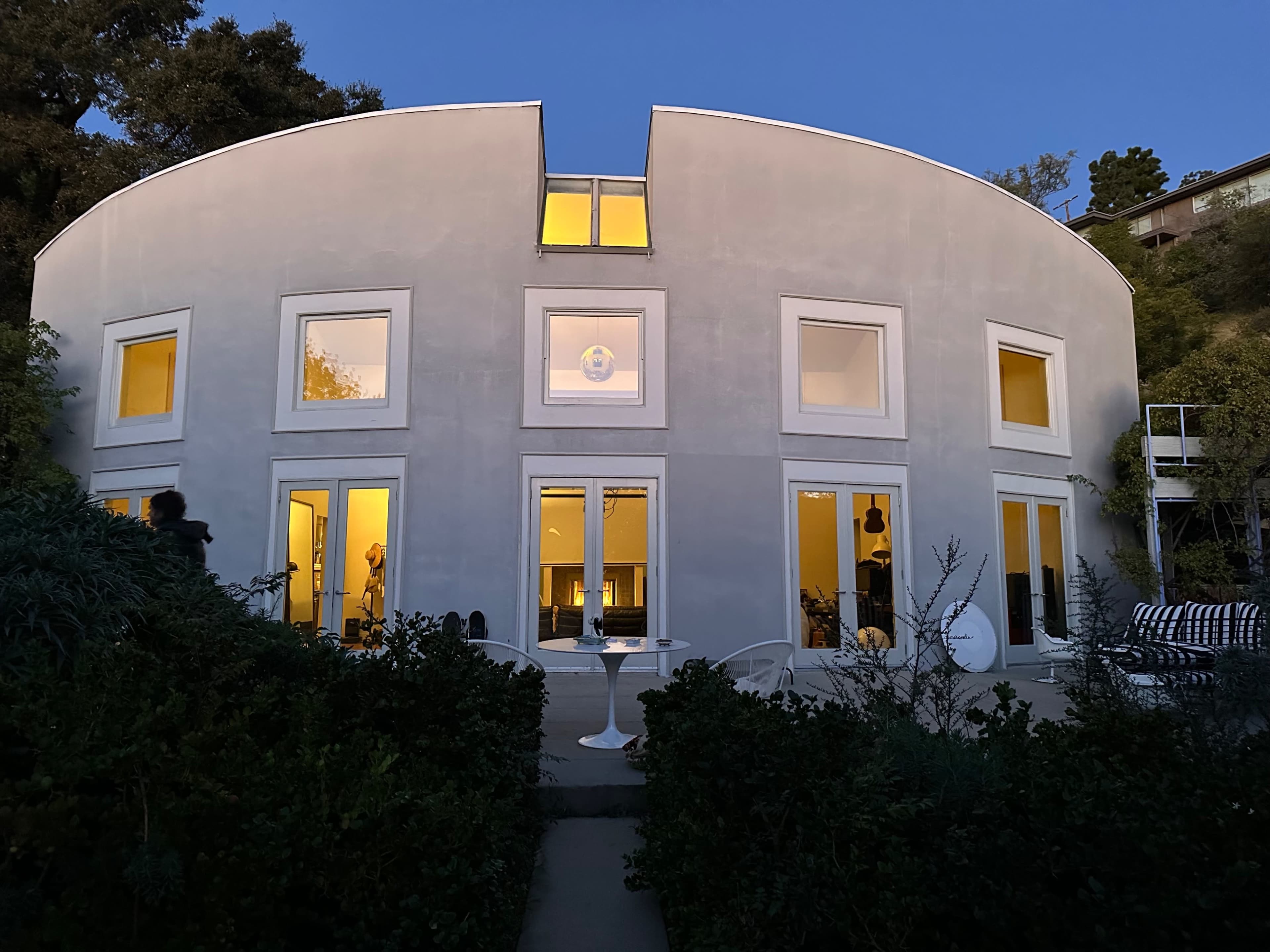The image shows a modern, rounded house with large windows illuminated at dusk, surrounded by greenery and outdoor furniture.