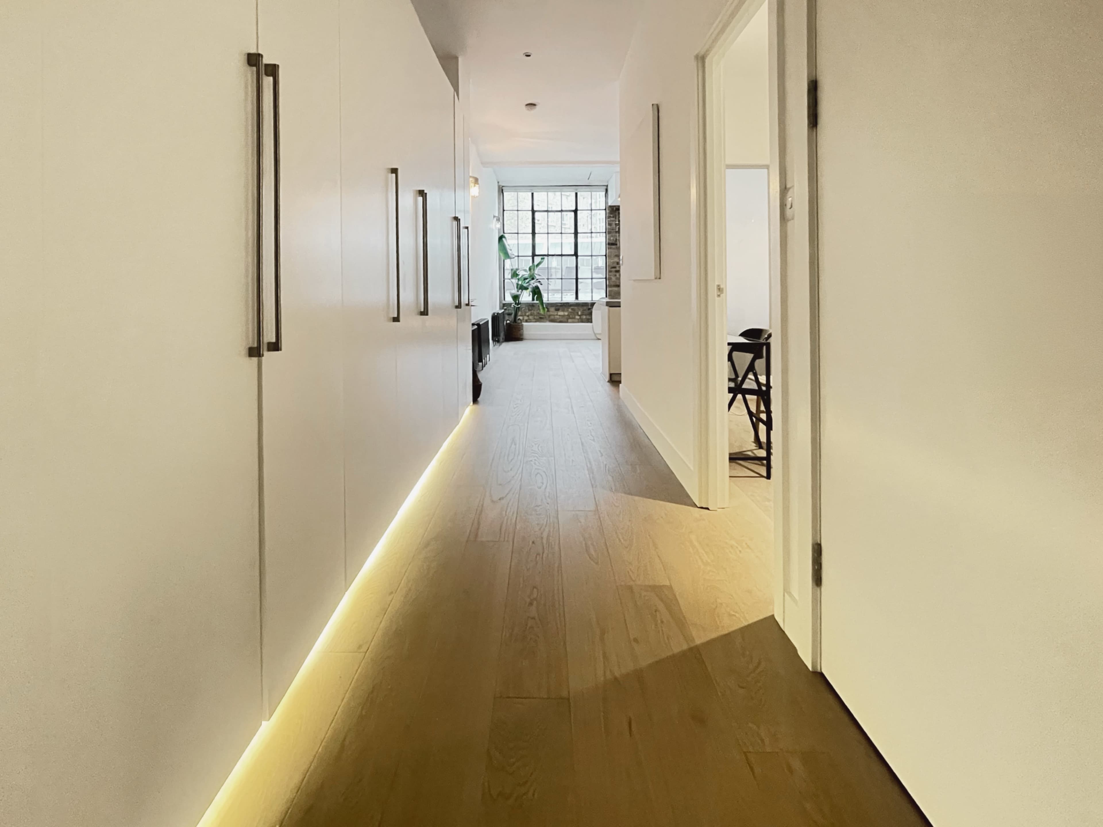 The image shows a well-lit corridor with white walls, wooden flooring, and cabinets on one side, leading to a room with a large window at the end.
