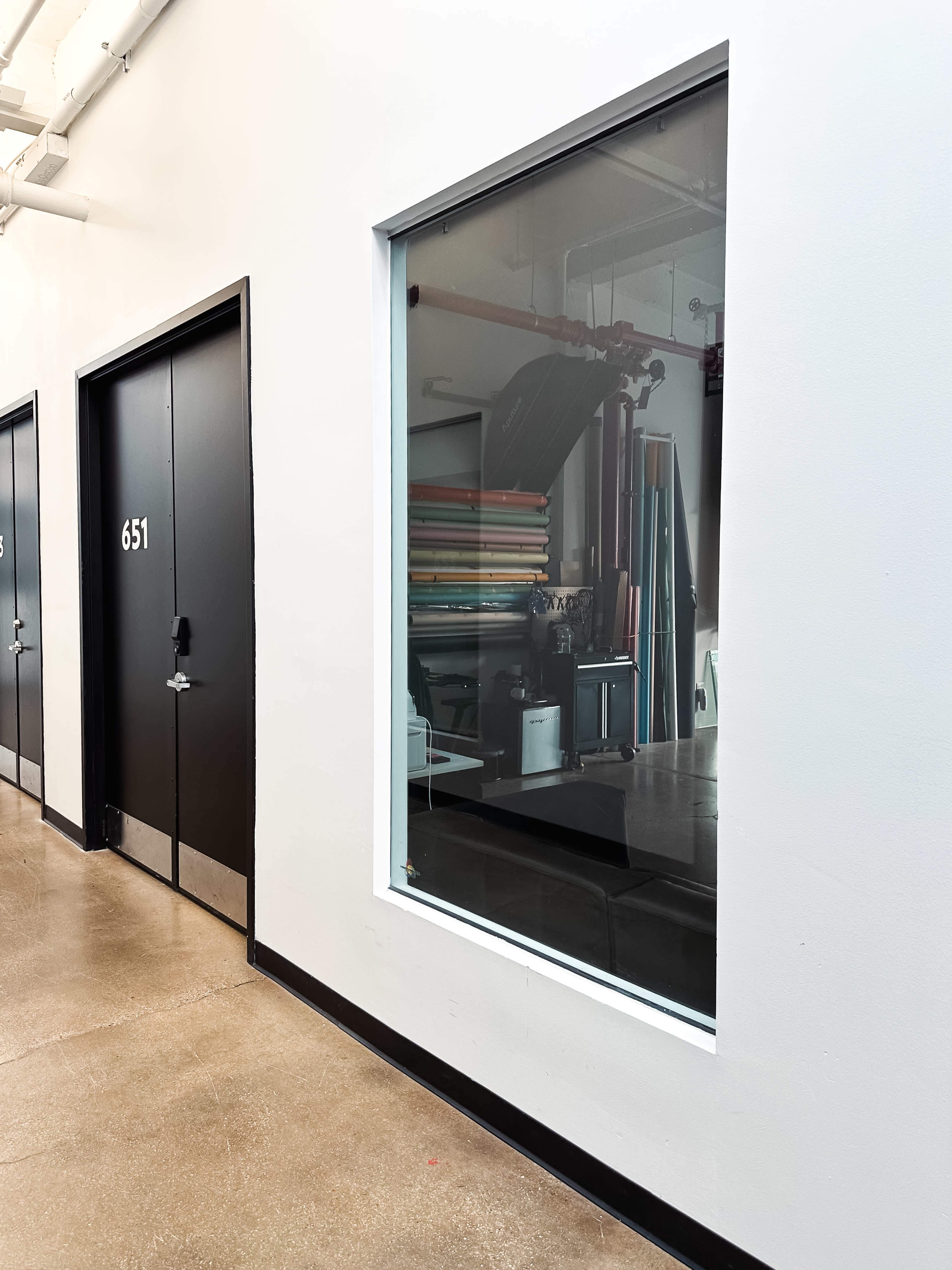 A corridor features a large window showing an interior space with various colored materials and equipment.