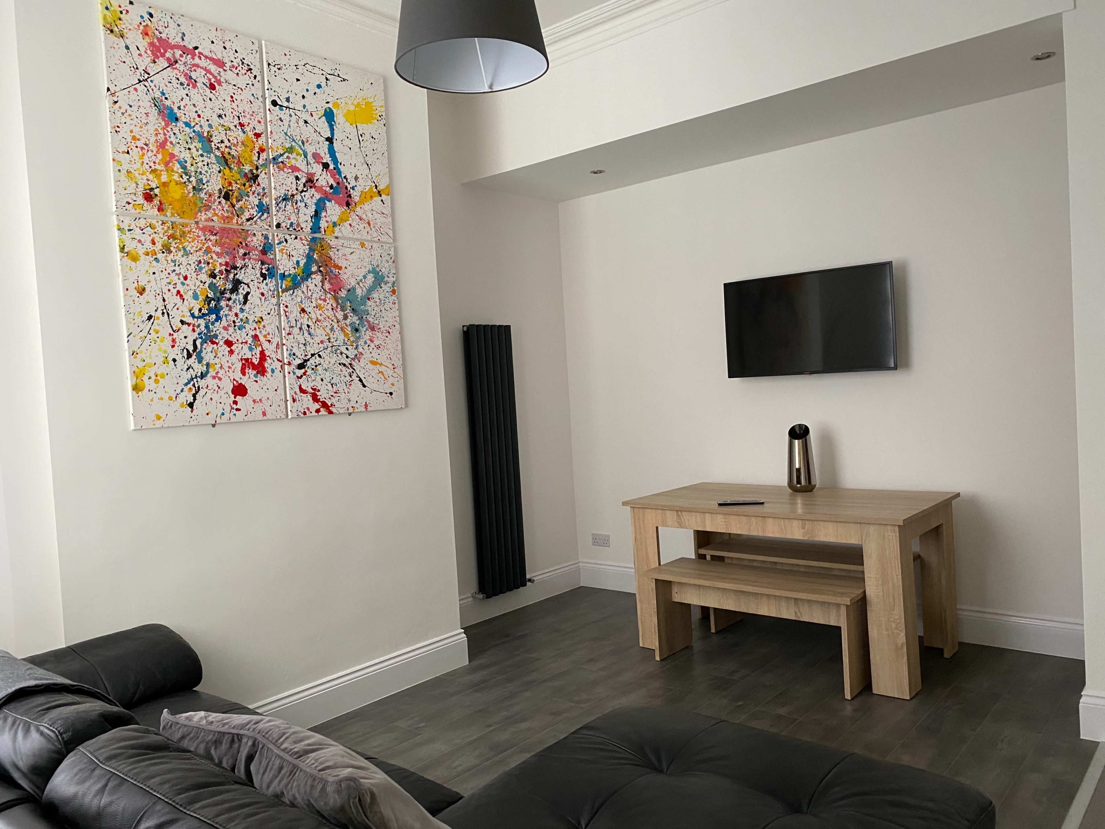 The room features a simple wooden table, a wall-mounted television, a large abstract painting, and a black sofa.