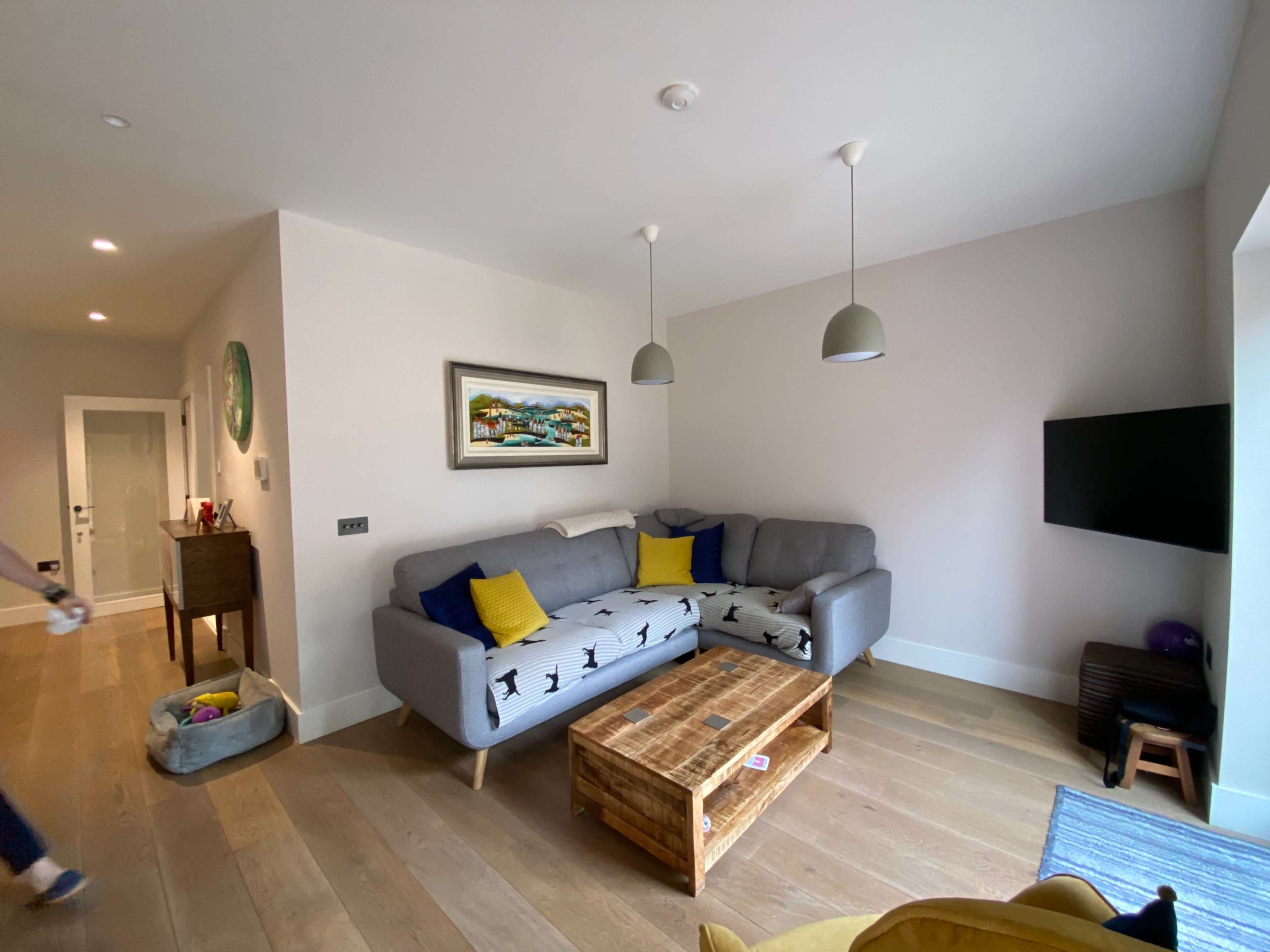 The room features a gray sofa with yellow cushions, a wooden coffee table, and a mounted television against a light-colored wall.