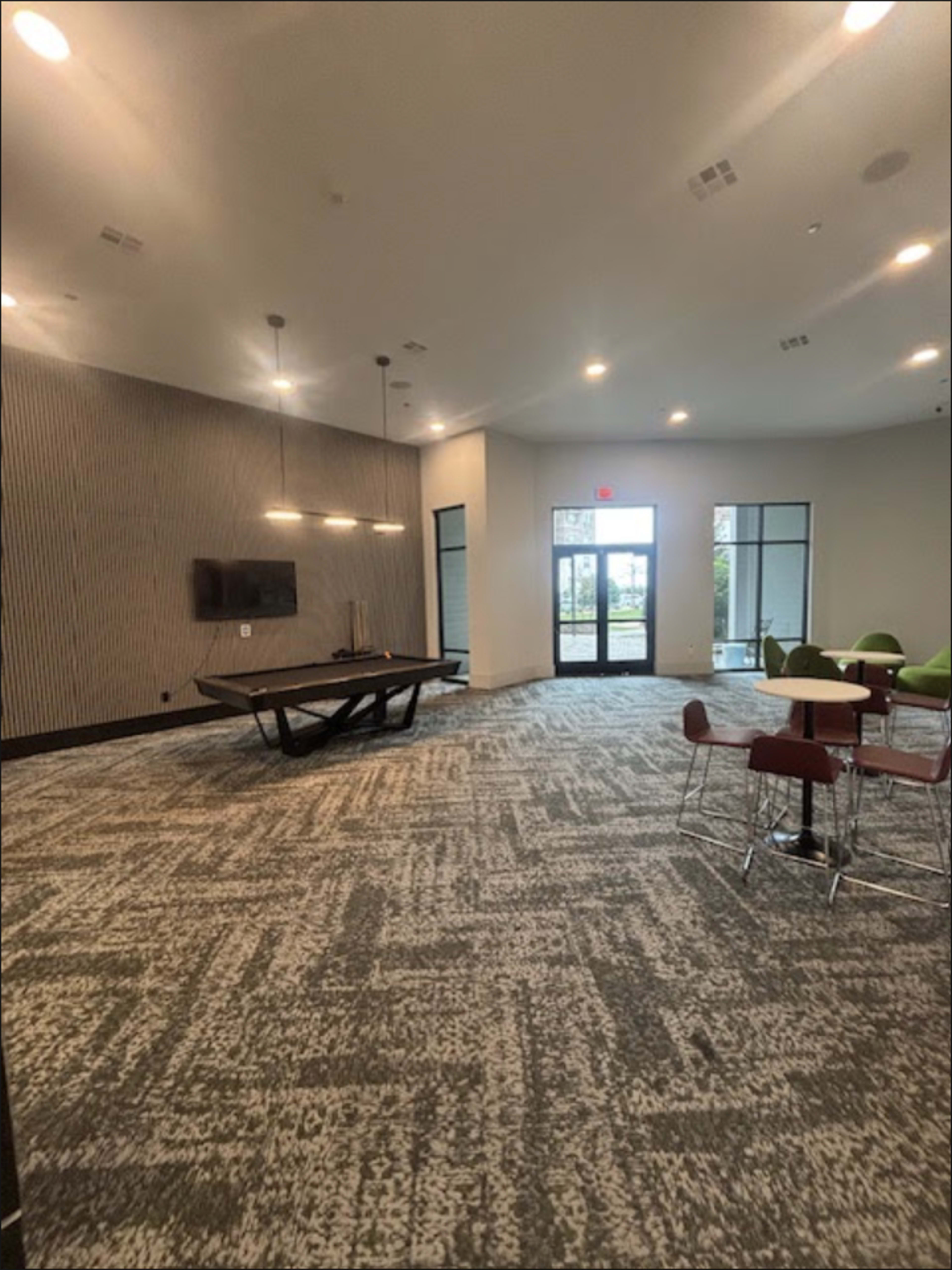 The image shows a spacious indoor lounge area featuring a billiards table, chairs, and large windows that let in natural light.