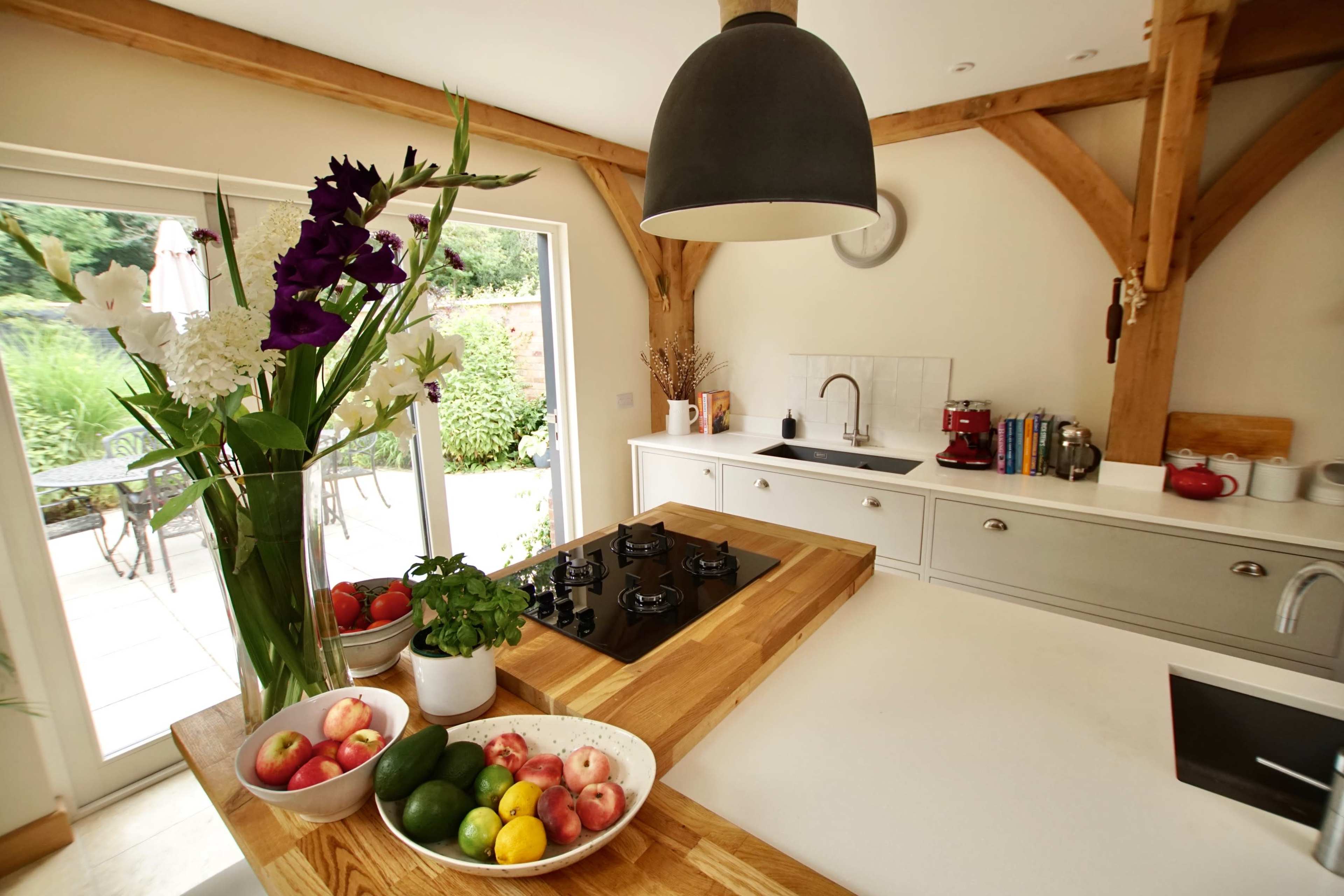 A modern kitchen features a wooden island with a bowl of fruit, a gas stovetop, and large windows looking into a garden.
