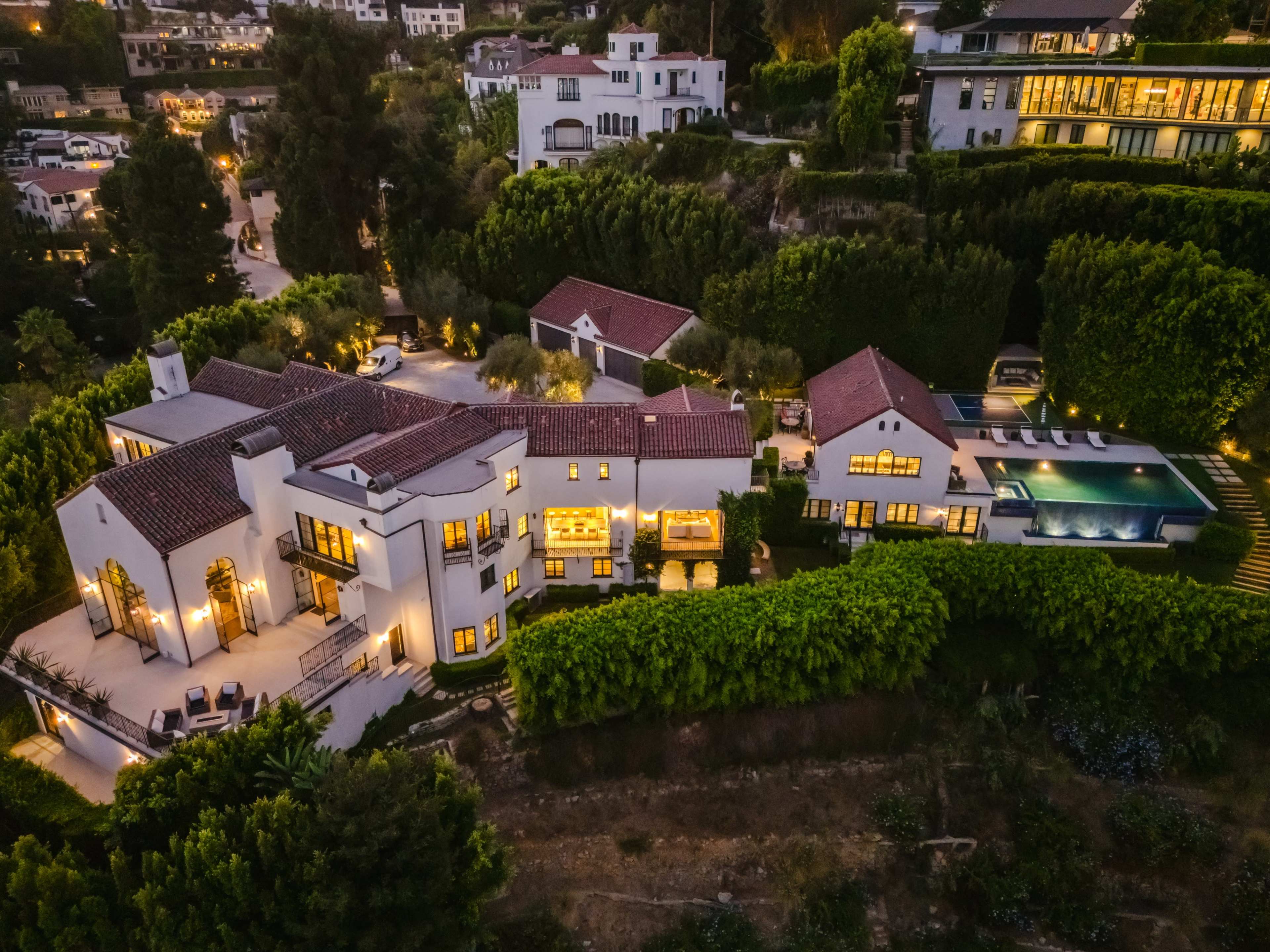 The image shows an aerial view of a sprawling luxury home with a swimming pool, surrounded by lush greenery and landscaped gardens in a hillside neighborhood.