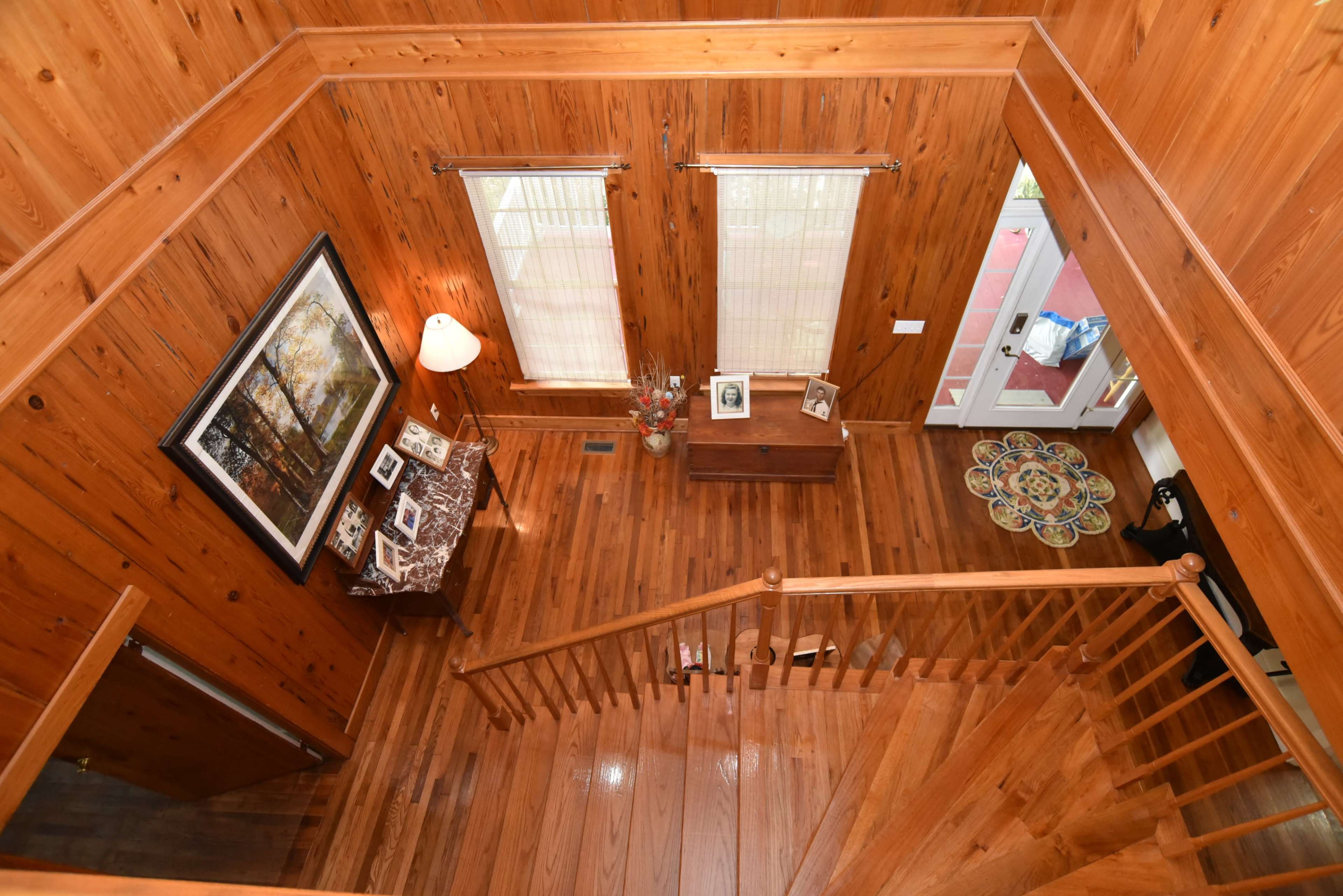 The image shows an interior view of a wooden-finished foyer with a staircase leading down, framed artwork on the wall, and a decorative rug on the floor.