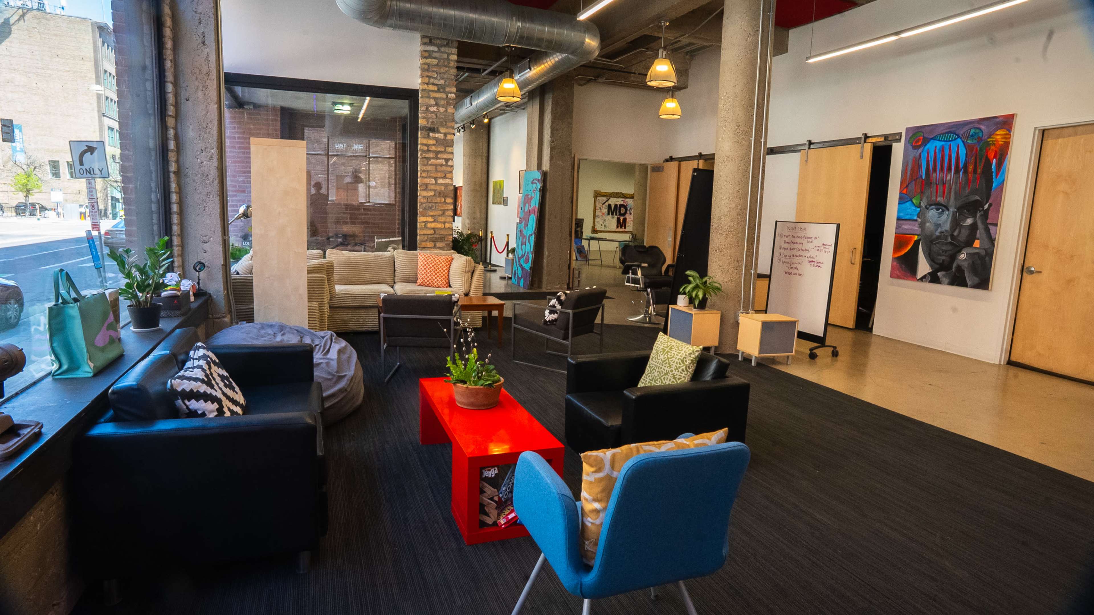The image depicts a modern, open lounge area with several seating options, including chairs and couches, surrounded by large windows and artwork on the walls.