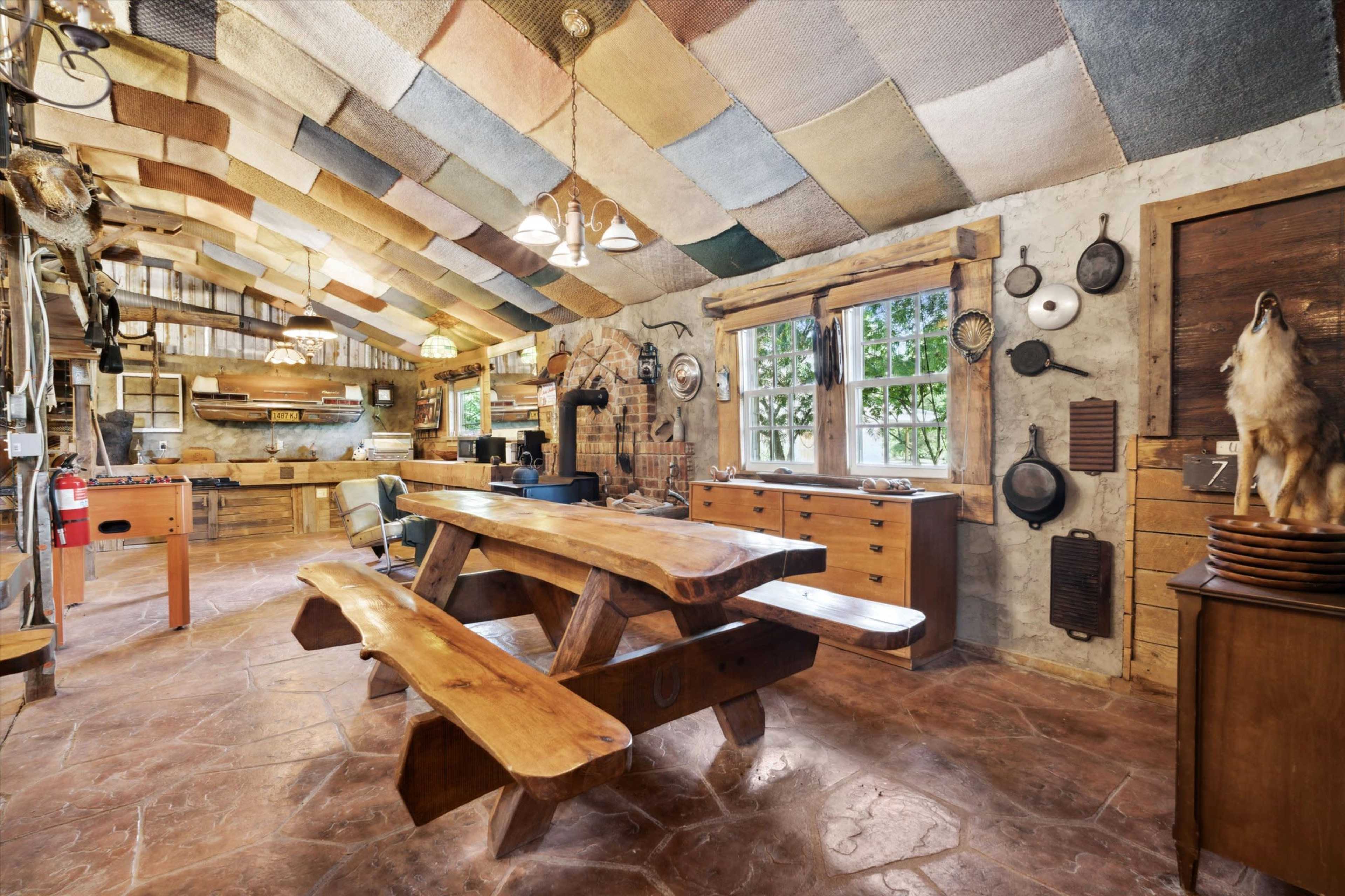 The image shows a rustic kitchen space featuring a large wooden dining table, stone floor, and a ceiling adorned with colorful textile panels.