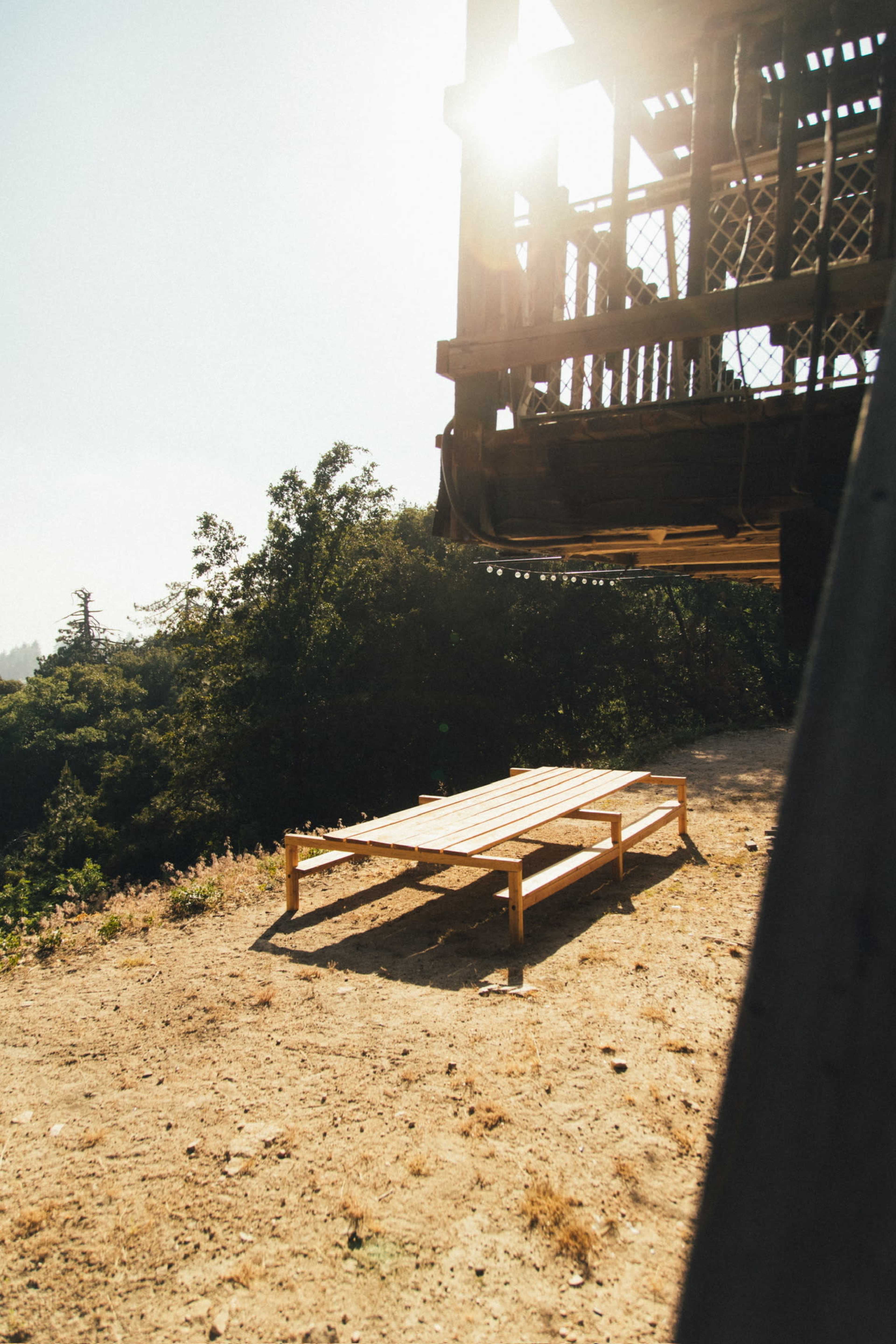 A wooden platform stands on a dirt area beneath a raised structure, with sunlight filtering through nearby trees.