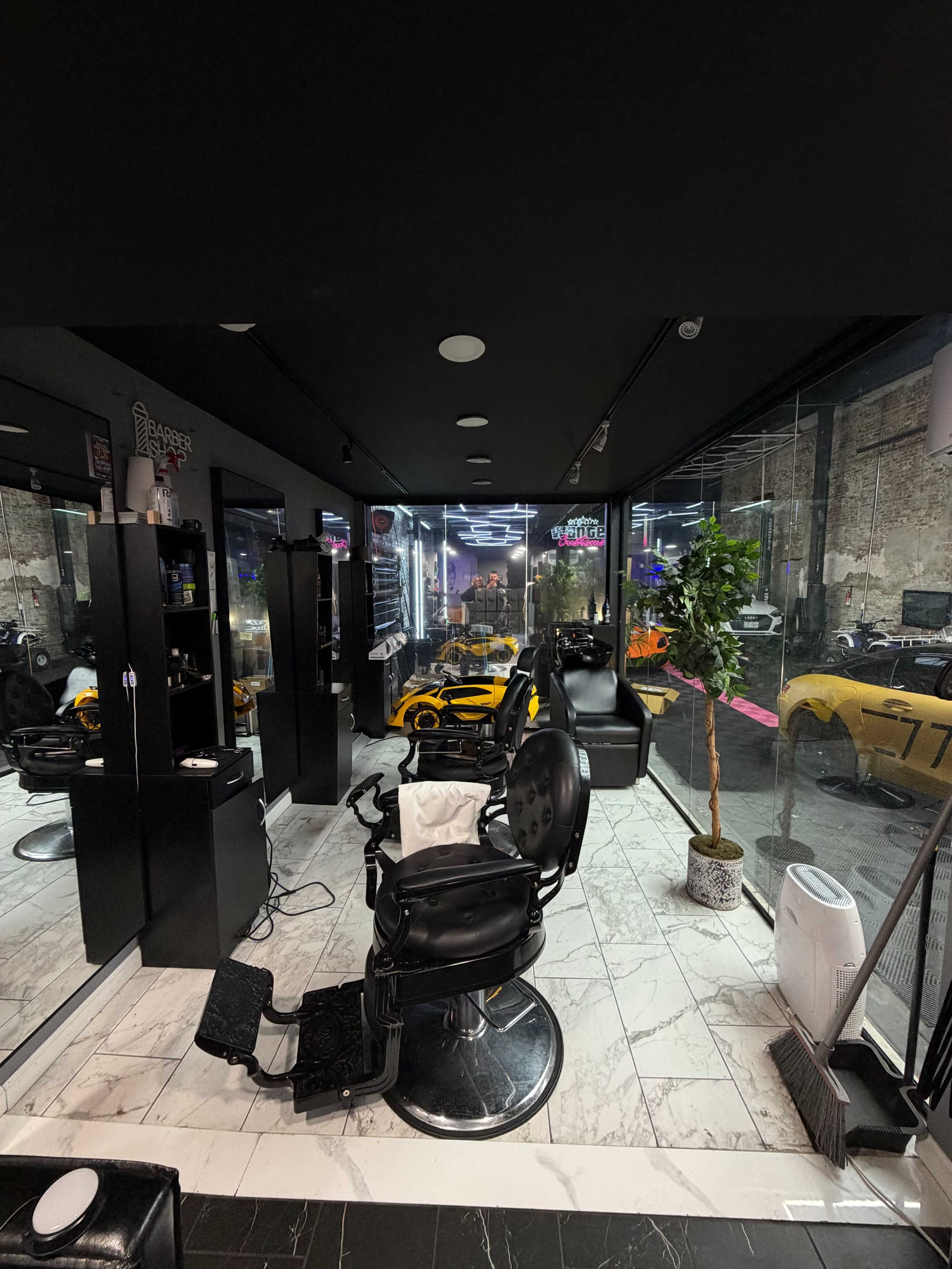 The image shows a modern barber shop with black walls, sleek chairs, mirrors, and a view of luxury cars through large windows.