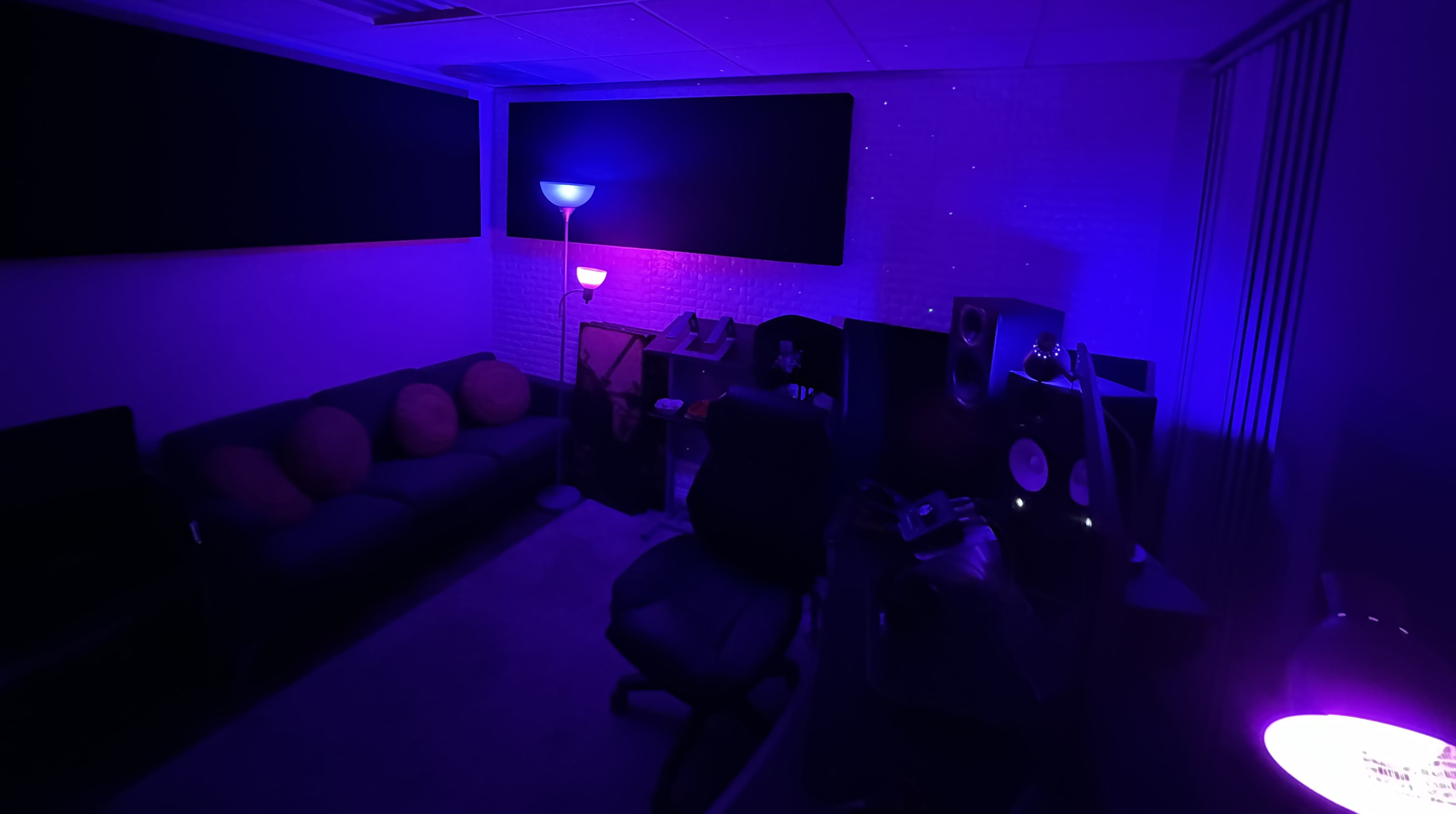 A dimly lit recording studio featuring a sofa, a desk with speakers, and a lamp emitting purple and blue light.