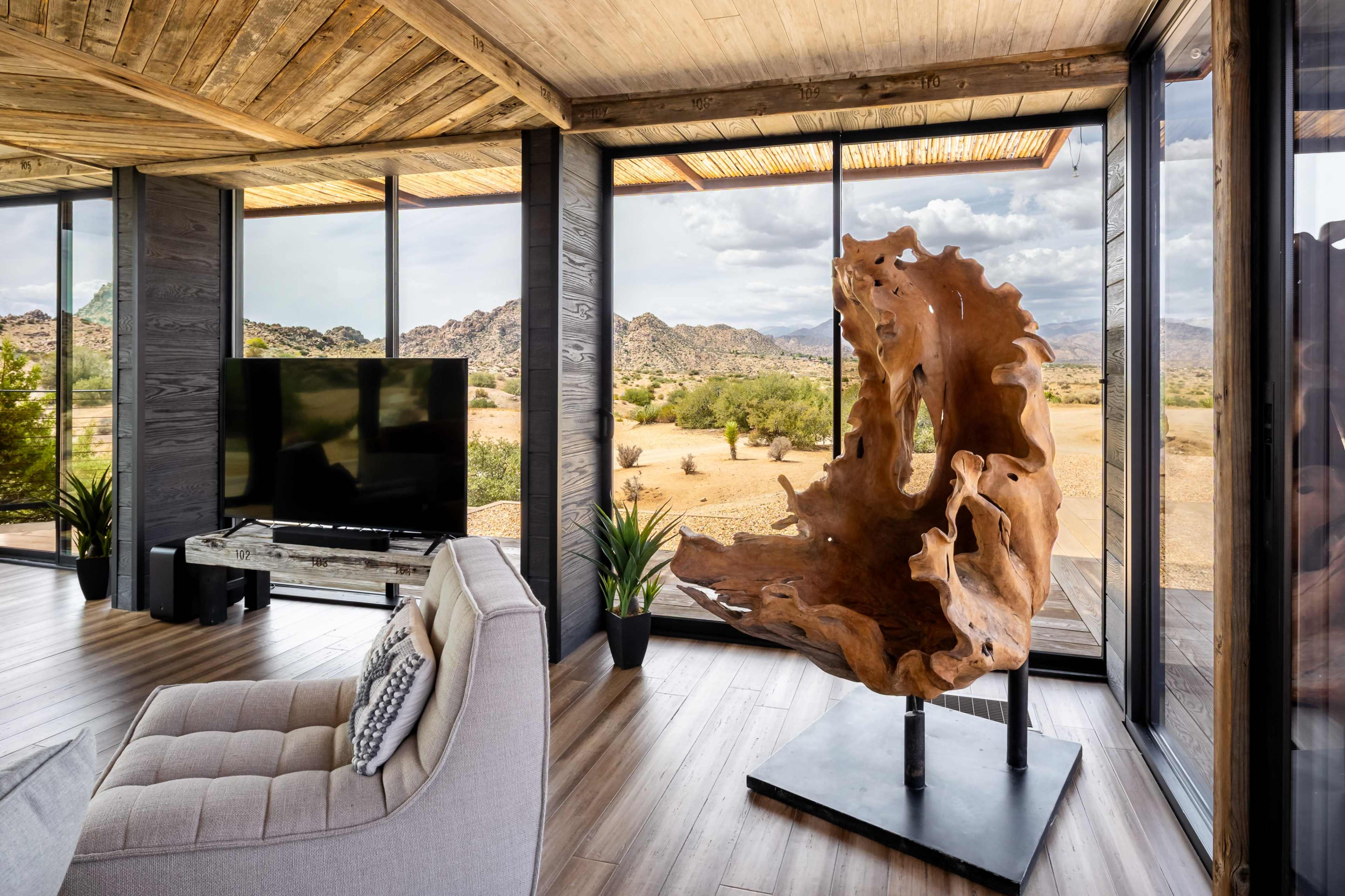 A modern living room features large windows with a view of a desert landscape, a television, and a unique wooden sculpture on display.