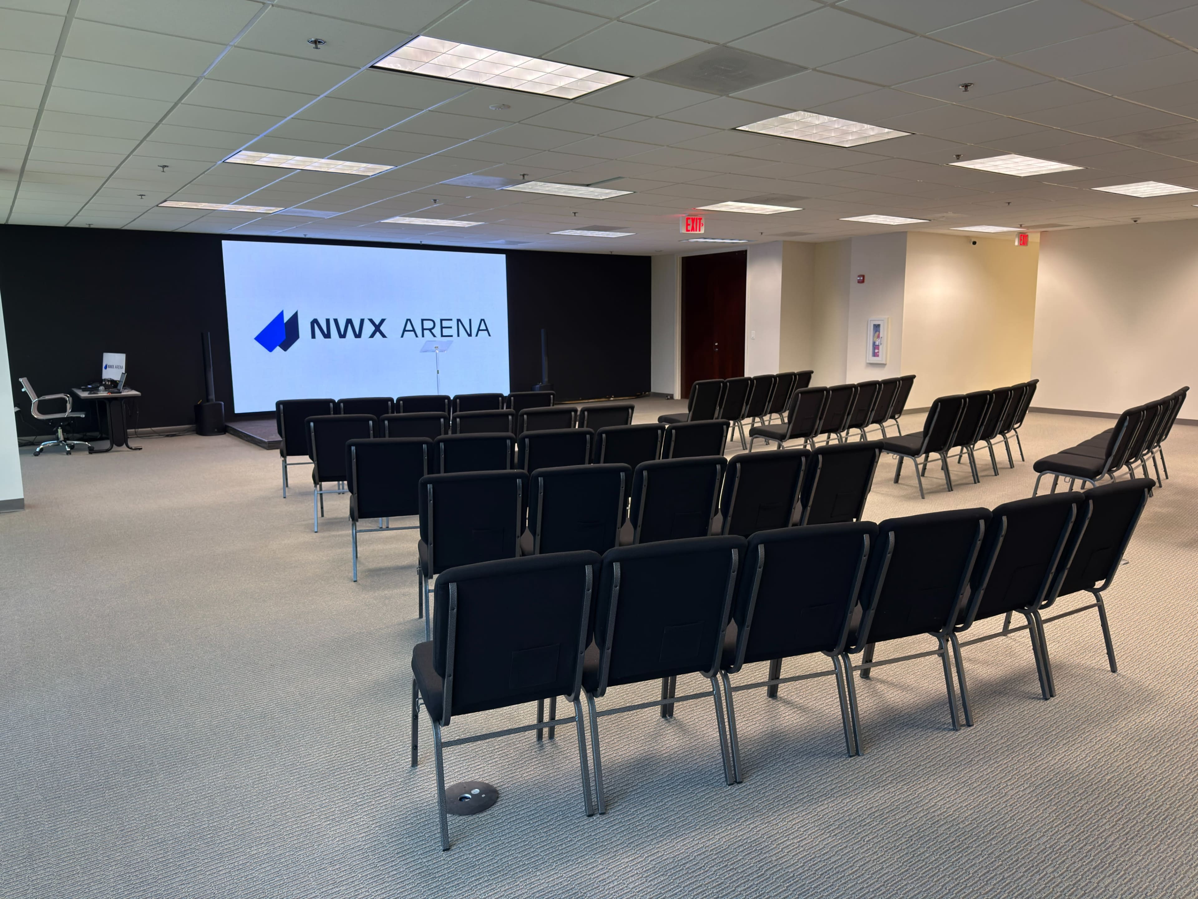 The image shows a conference room with rows of black chairs facing a large screen displaying the logo "NWX ARENA."