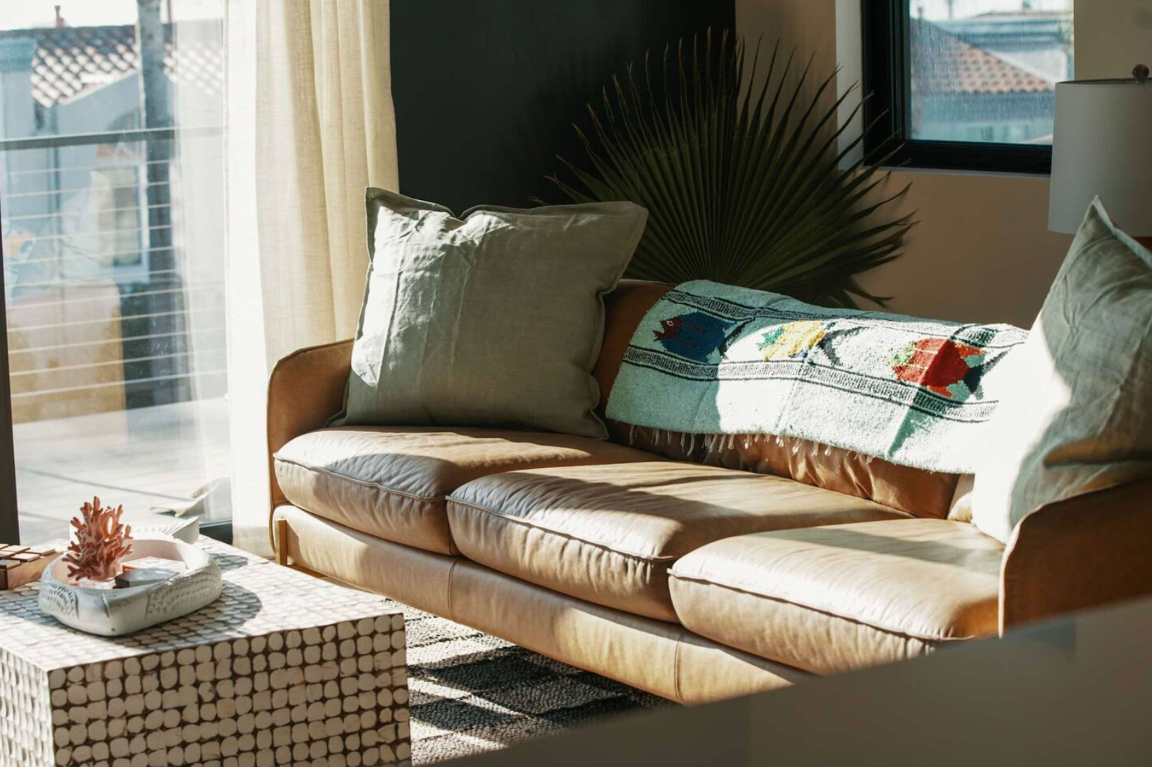 A brown leather sofa with decorative pillows sits beside a small coffee table in a sunlit room with large windows.