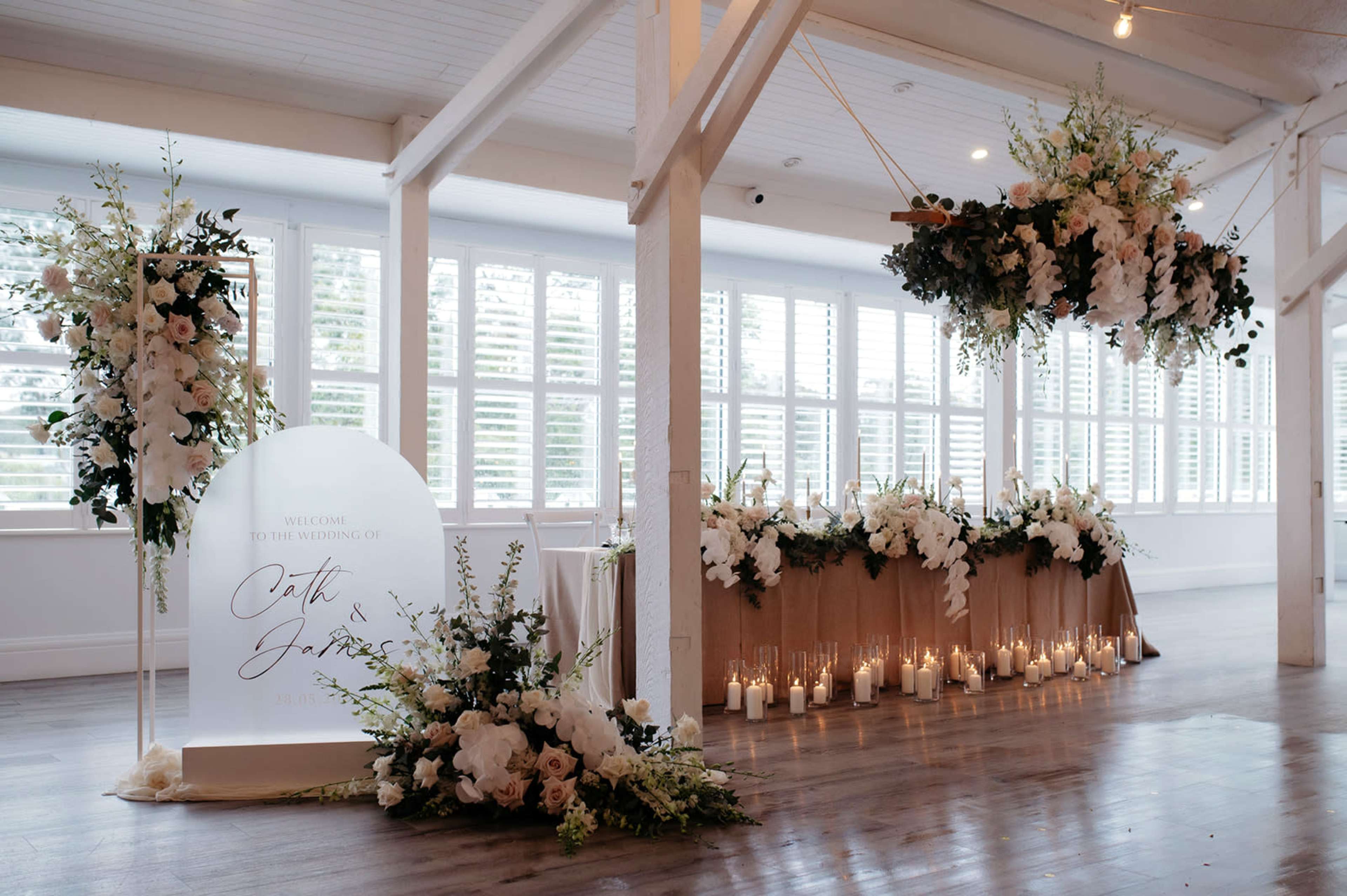 The venue features a decorated area for a wedding, including a floral arch, a long table adorned with flowers, and candles, all set under large windows.