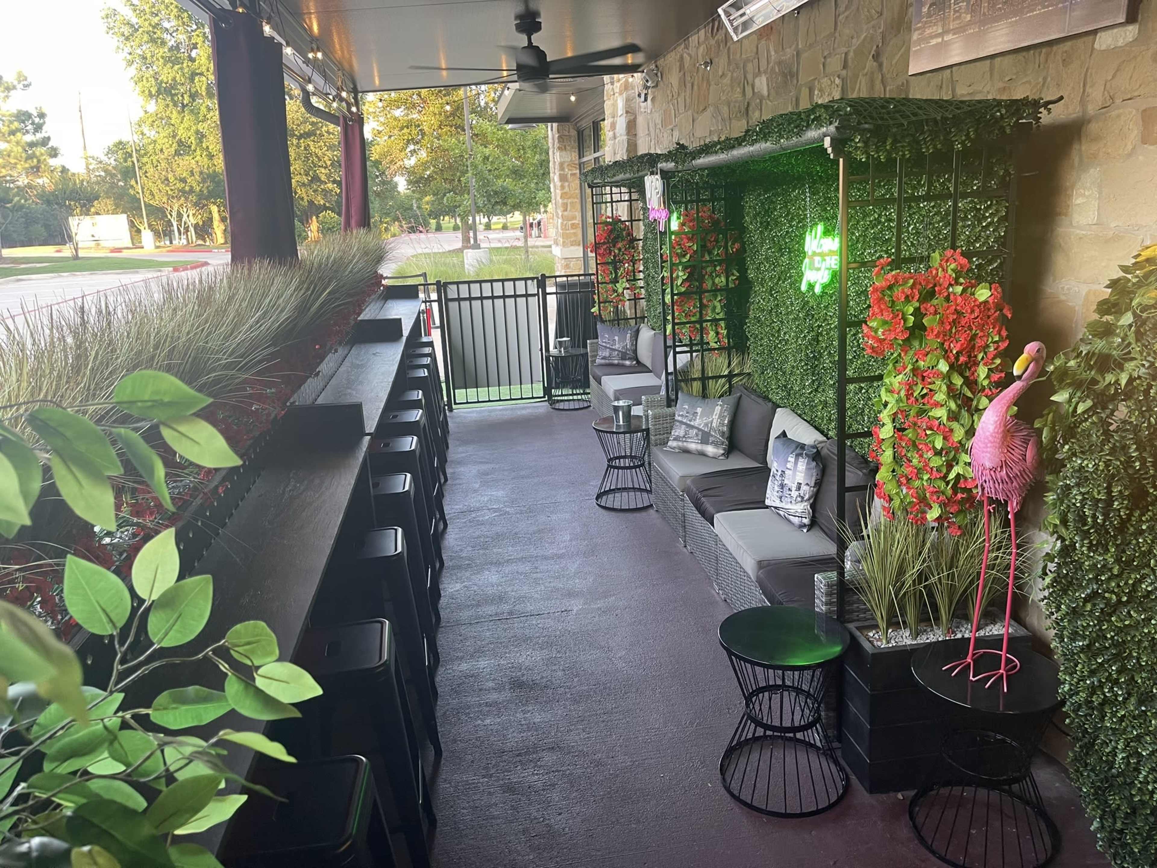 The image shows an outdoor seating area with stone walls, greenery, and decorative elements, including a pink flamingo.