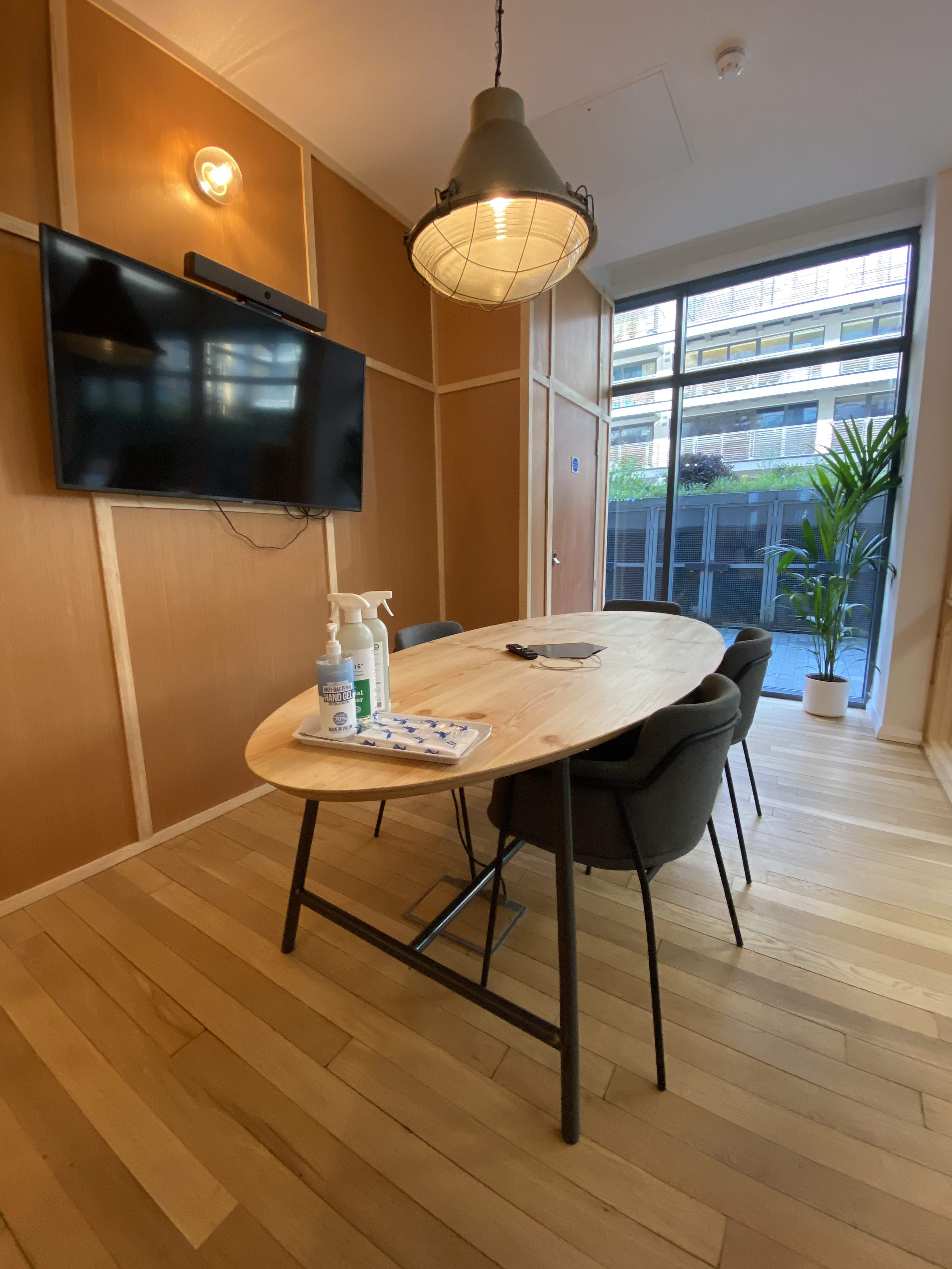 A modern meeting room features a wooden table with chairs, a wall-mounted TV, and a window overlooking greenery.