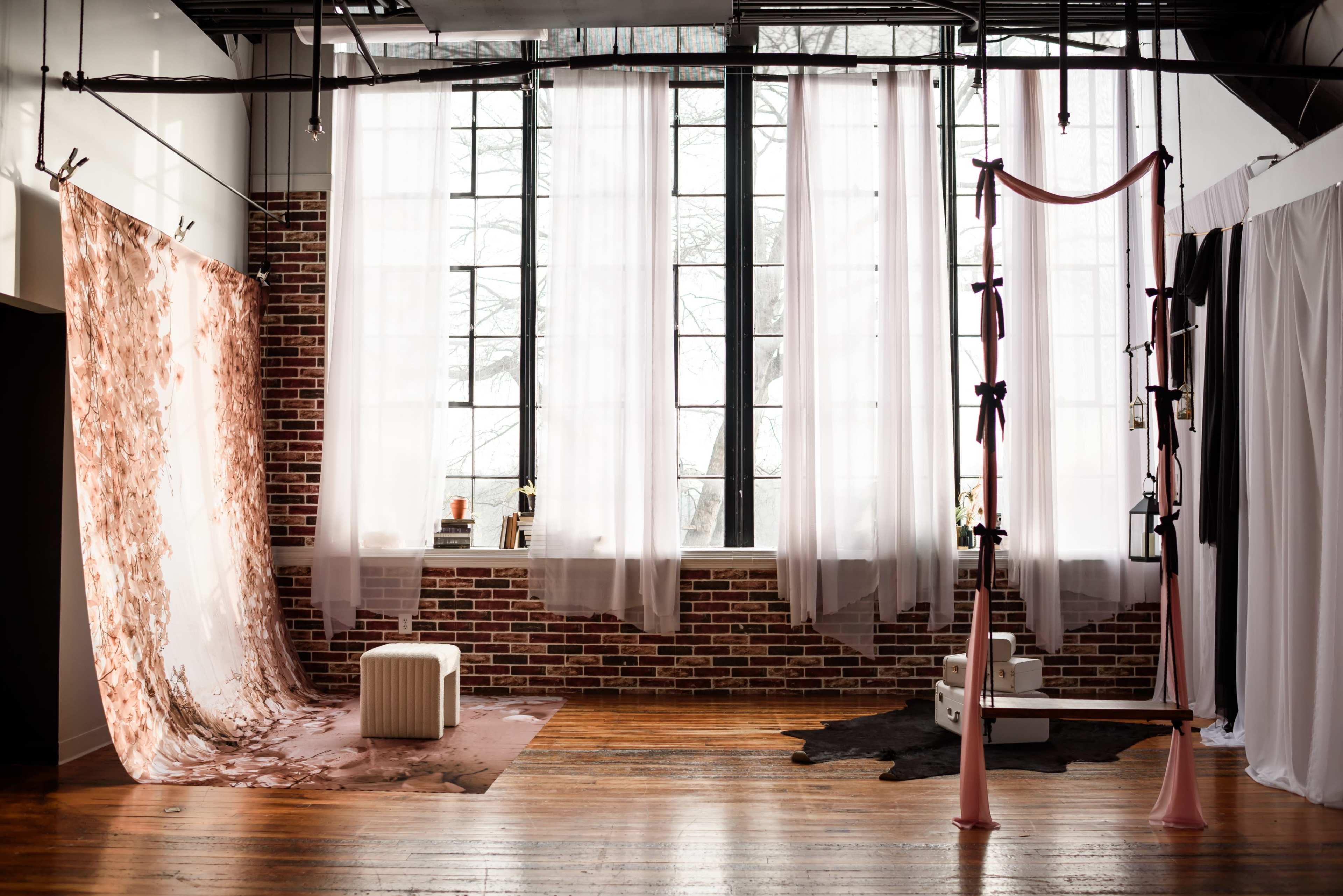 The image shows a spacious studio with large windows, featuring a combination of fabric backdrops, a wooden floor, and a Pilates reformer.