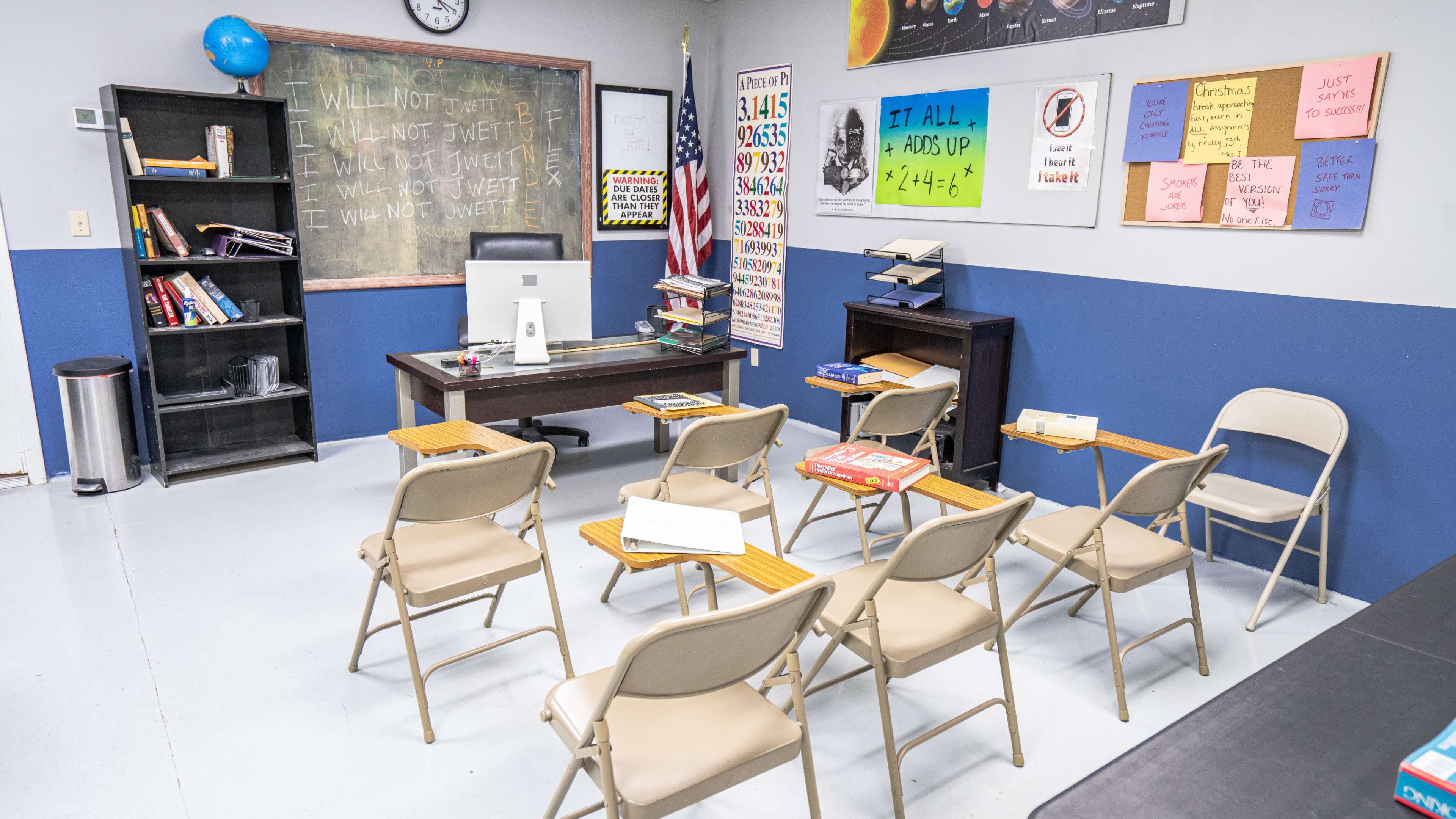 The image shows a classroom set up with a desk at the front, rows of chairs facing it, chalkboards on the walls, and various educational posters displayed.