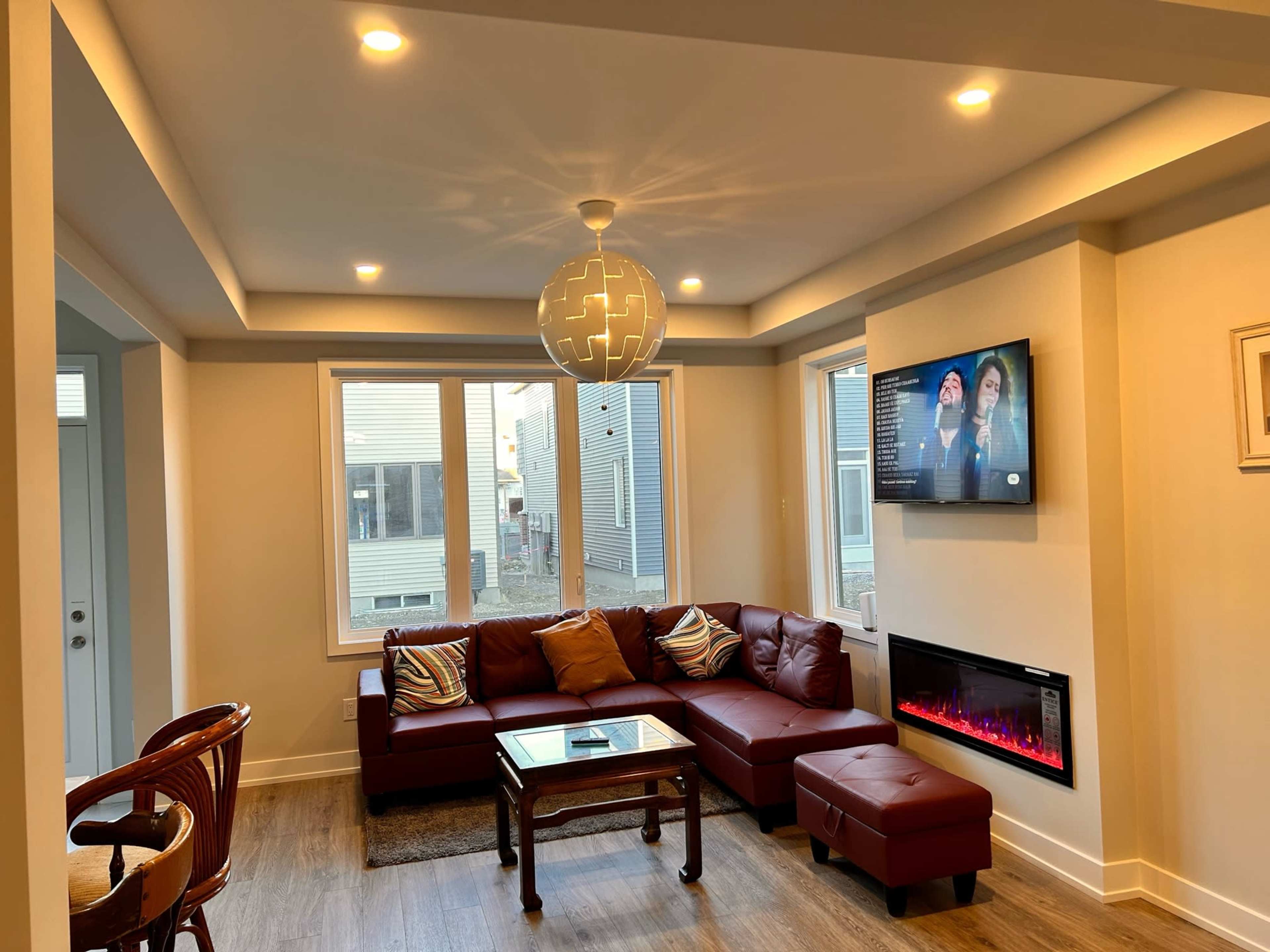 A modern living room features a brown leather sectional sofa, a glass coffee Table, a television on the wall, and an electric fireplace beneath it.