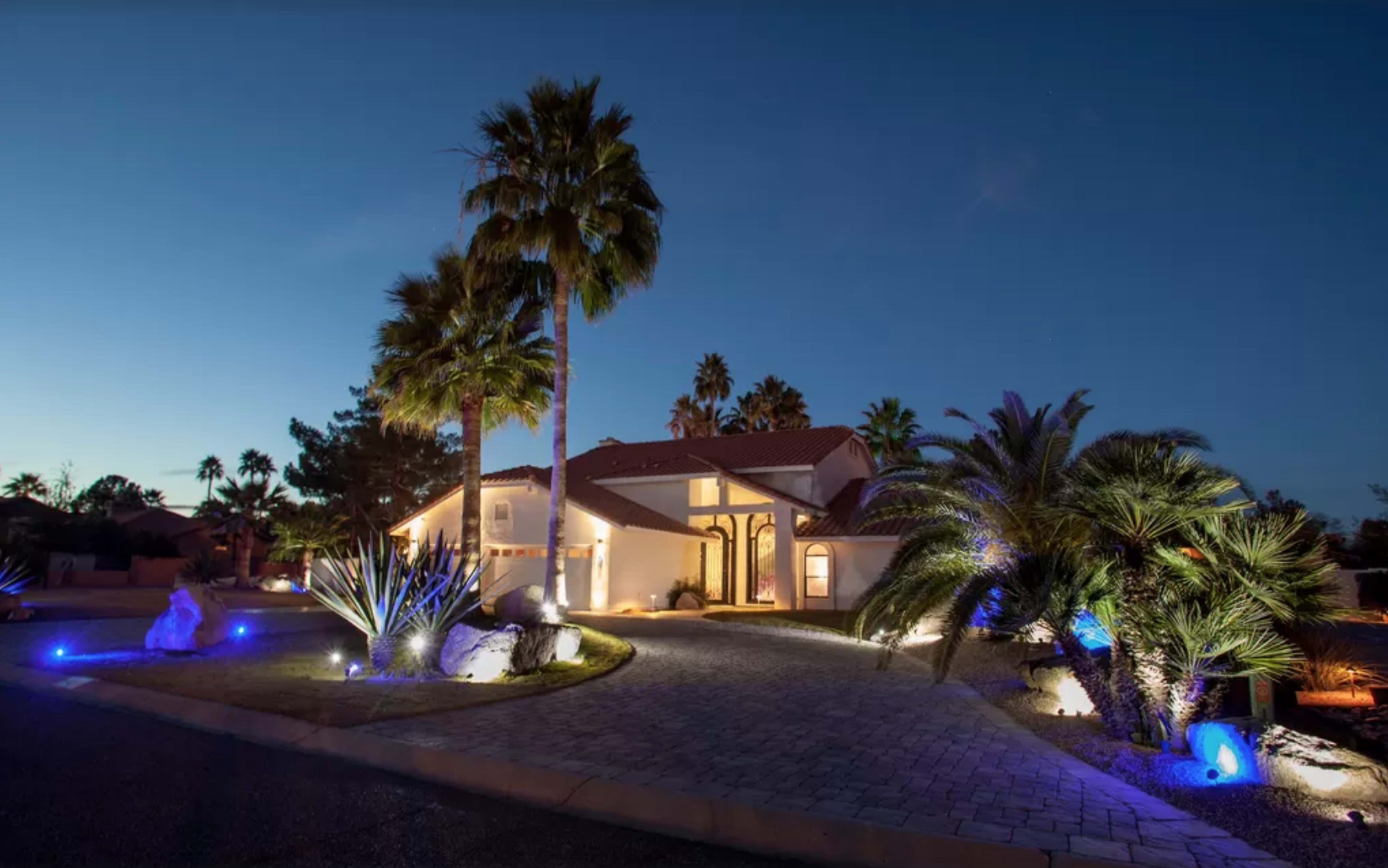 A modern house with a tiled roof is surrounded by palm trees and illuminated by landscape lighting at dusk.