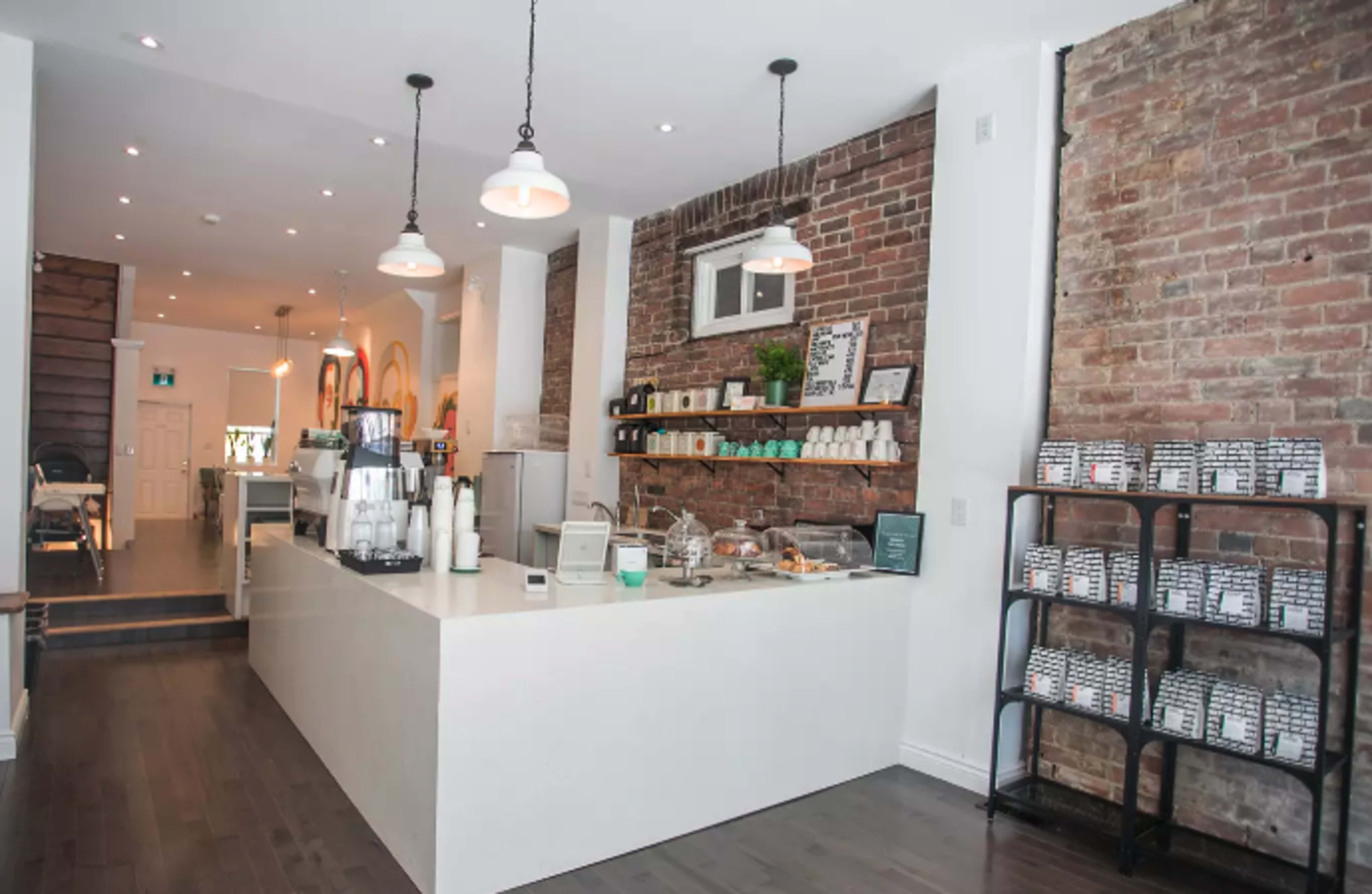 The image shows a modern cafe with a sleek white countertop, brick walls, and a display of shelves and seating areas in the background.