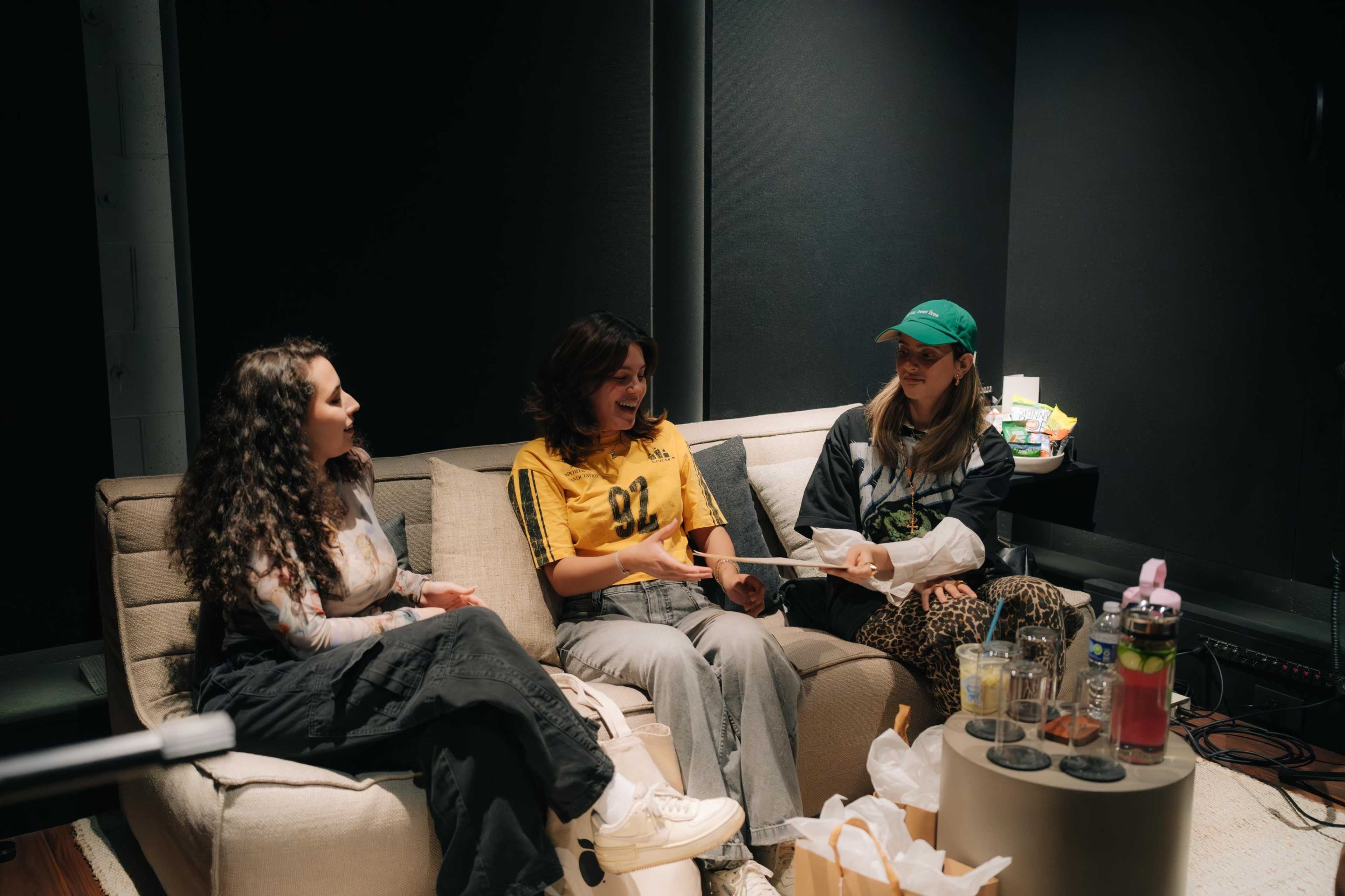 Three women relax on a couch in a dimly lit room, engaging in conversation with drinks and snacks on a nearby table.