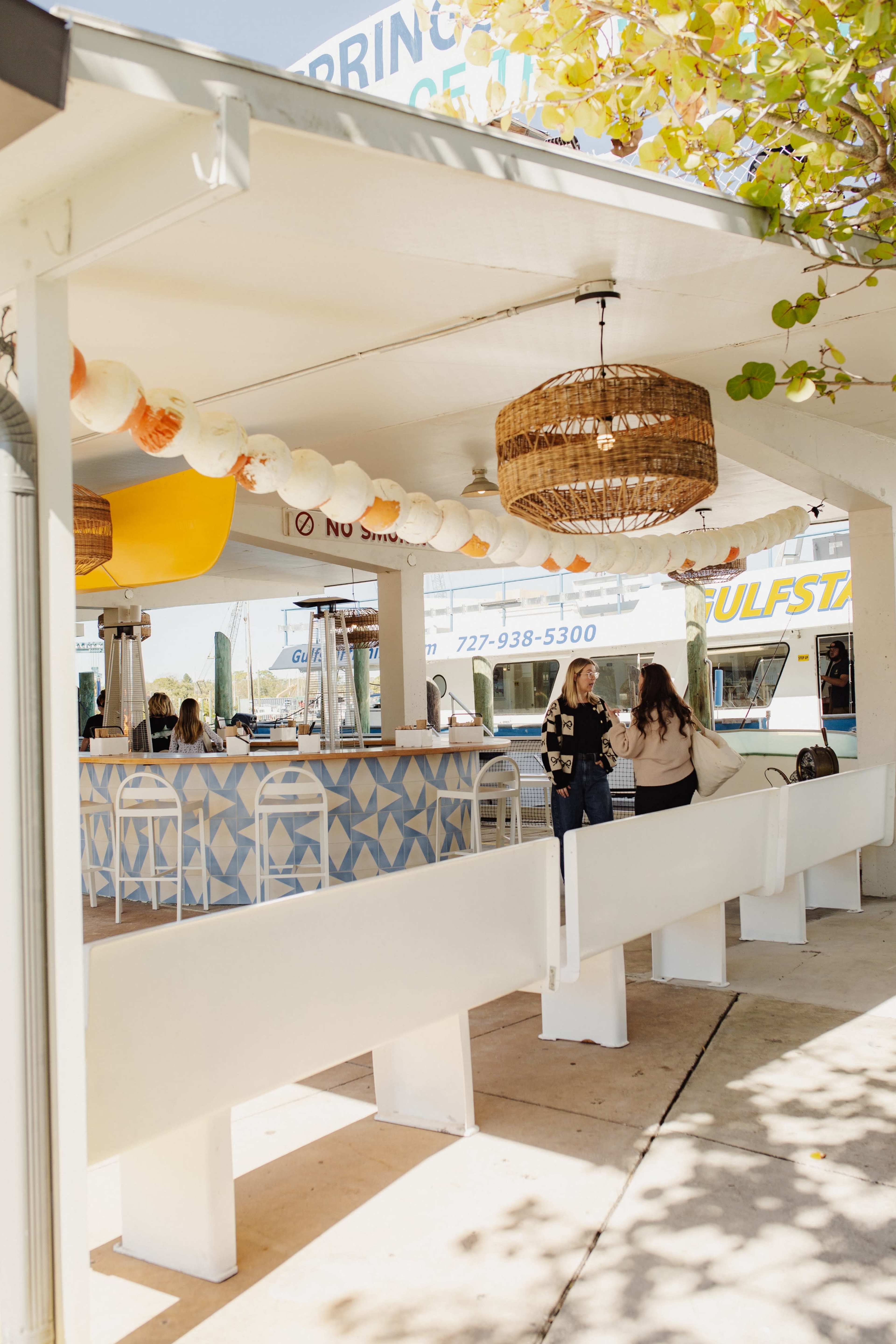 The image shows an outdoor food stall area with a patterned bar, hanging light fixtures, and patrons seated at the counter.