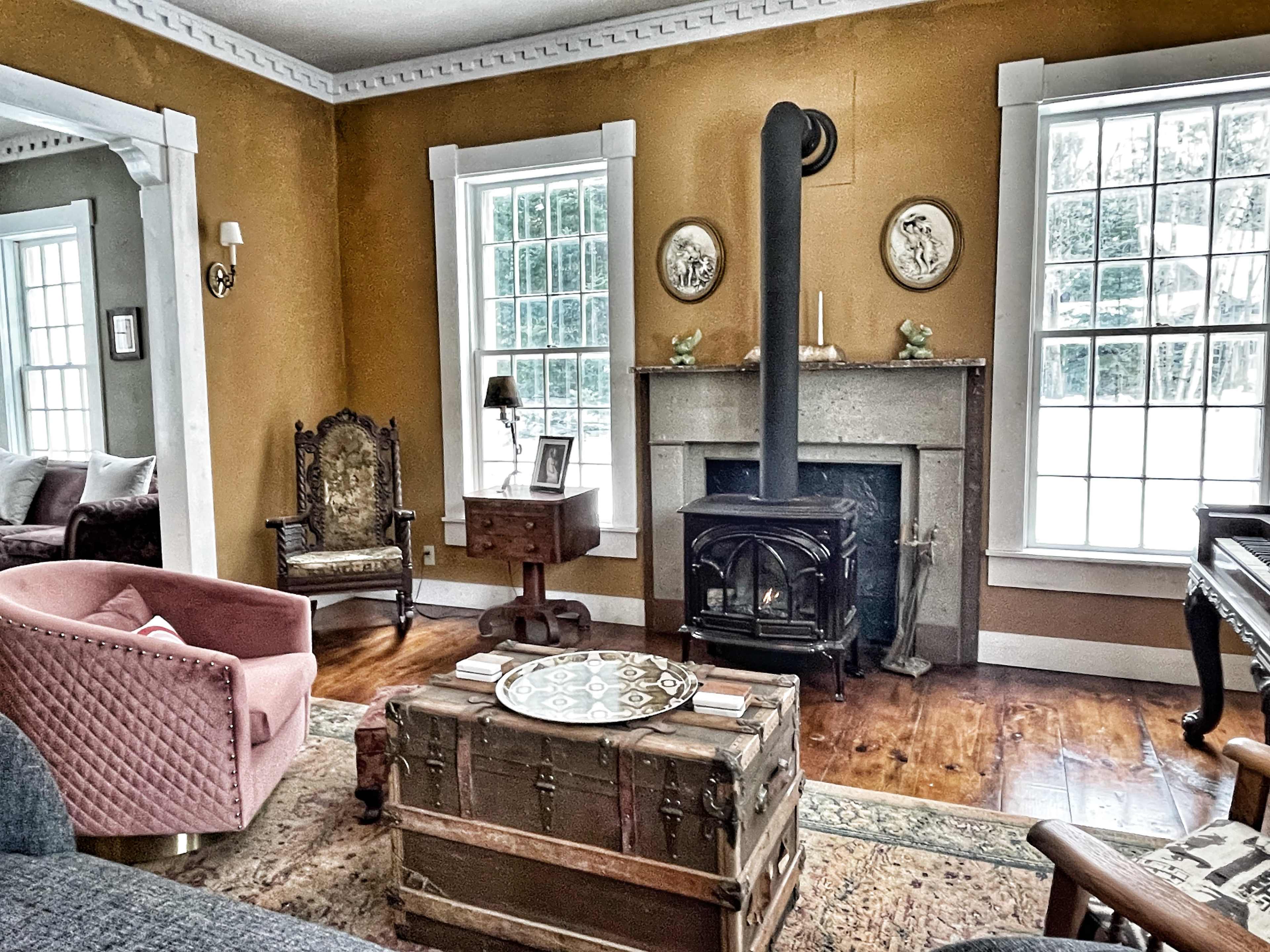 A cozy living room featuring a fireplace, vintage furniture, and large windows.