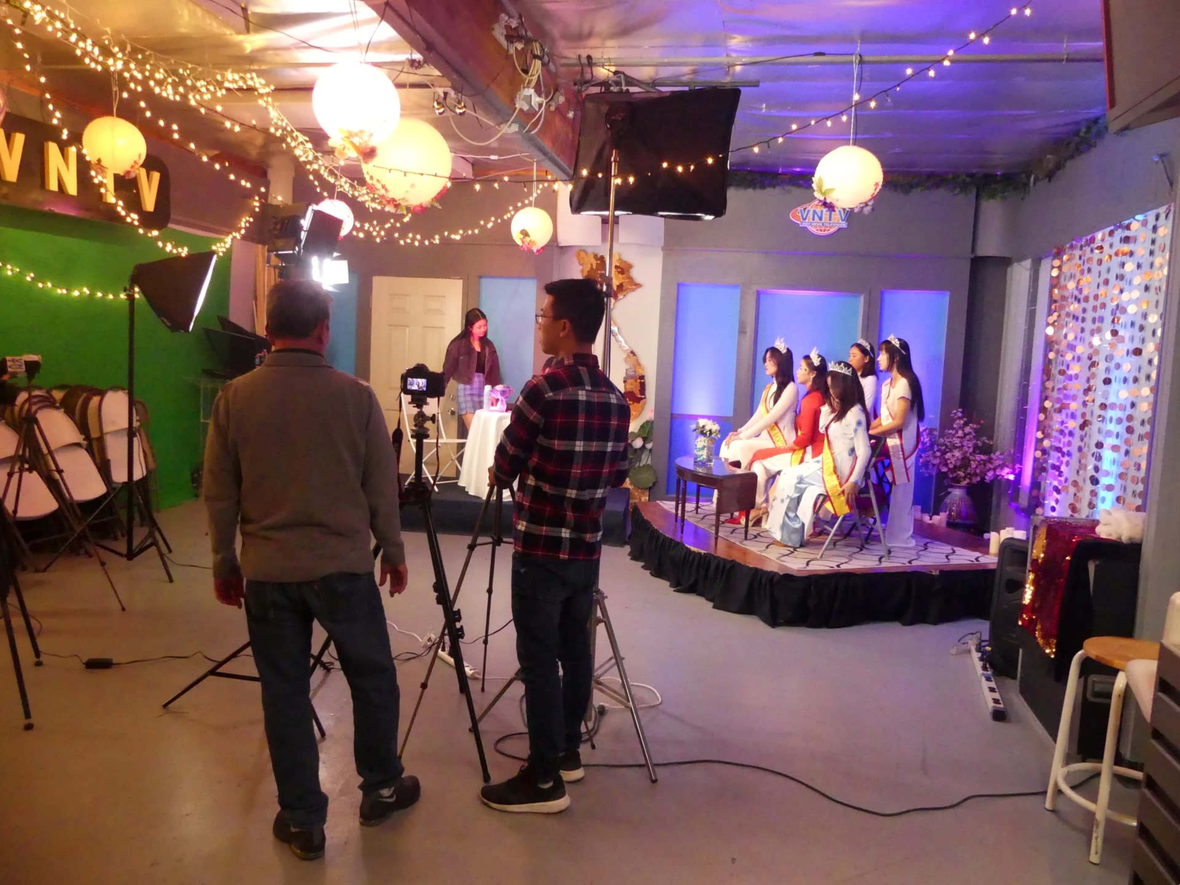 A production crew sets up lights and cameras for a staged performance featuring several seated performers in a decorated studio.