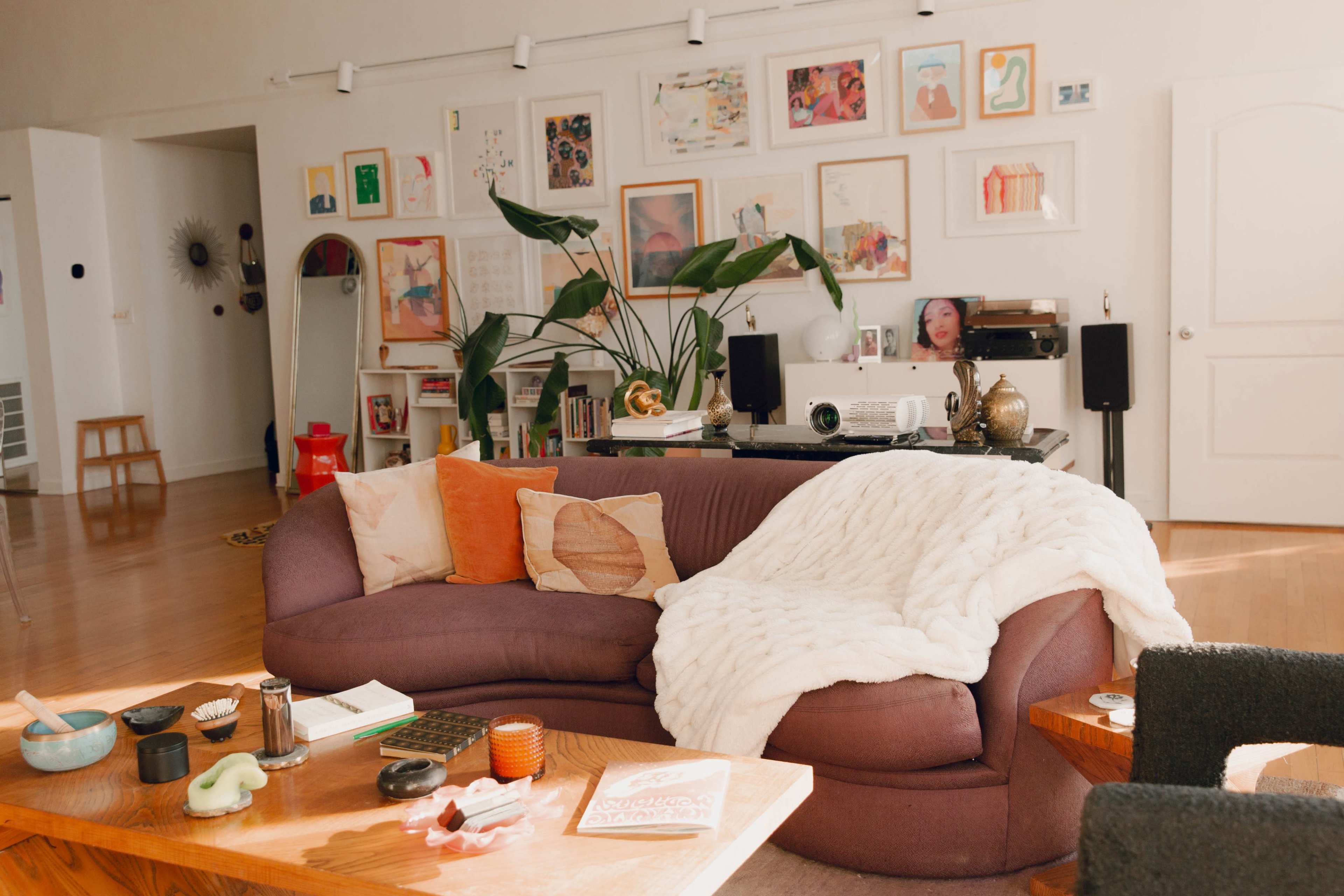 A cozy living room features a curved sofa with a throw blanket, surrounded by various art pieces on the walls and decor items on wooden tables.