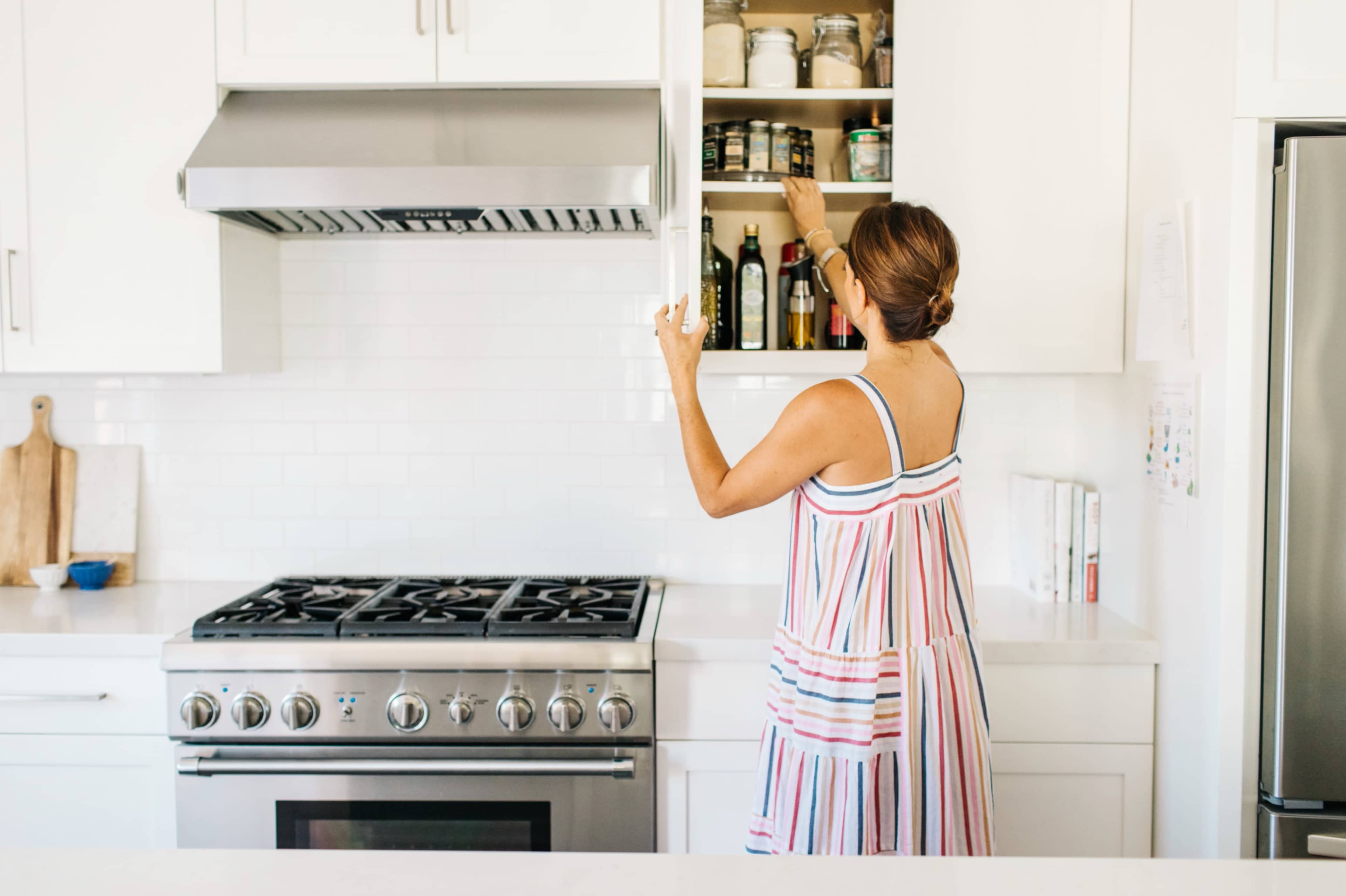 A person is reaching for a jar on a shelf in a modern kitchen with white cabinetry and a stainless steel stove.