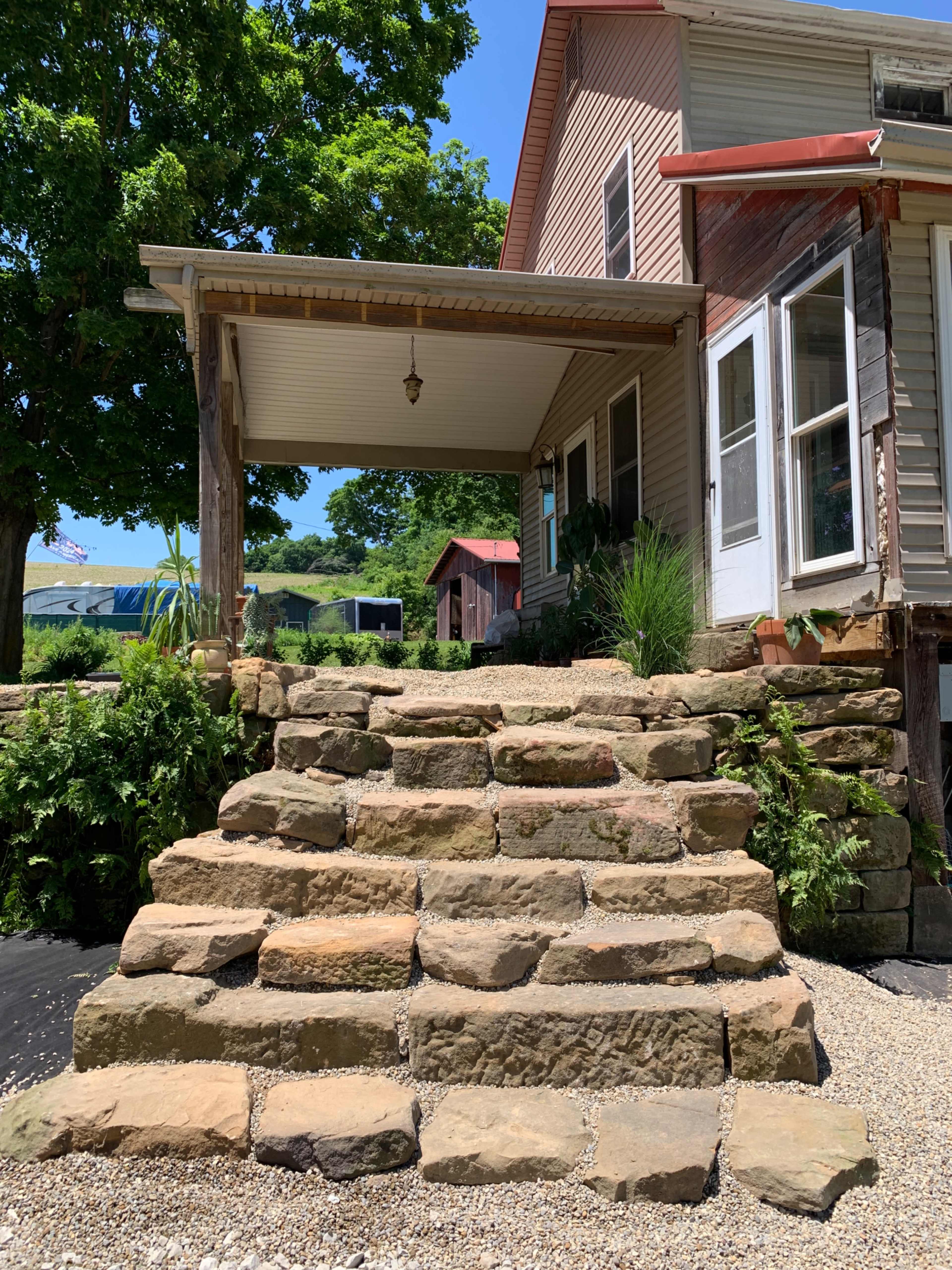 A set of stone steps leading up to a porch attached to a house, surrounded by greenery.