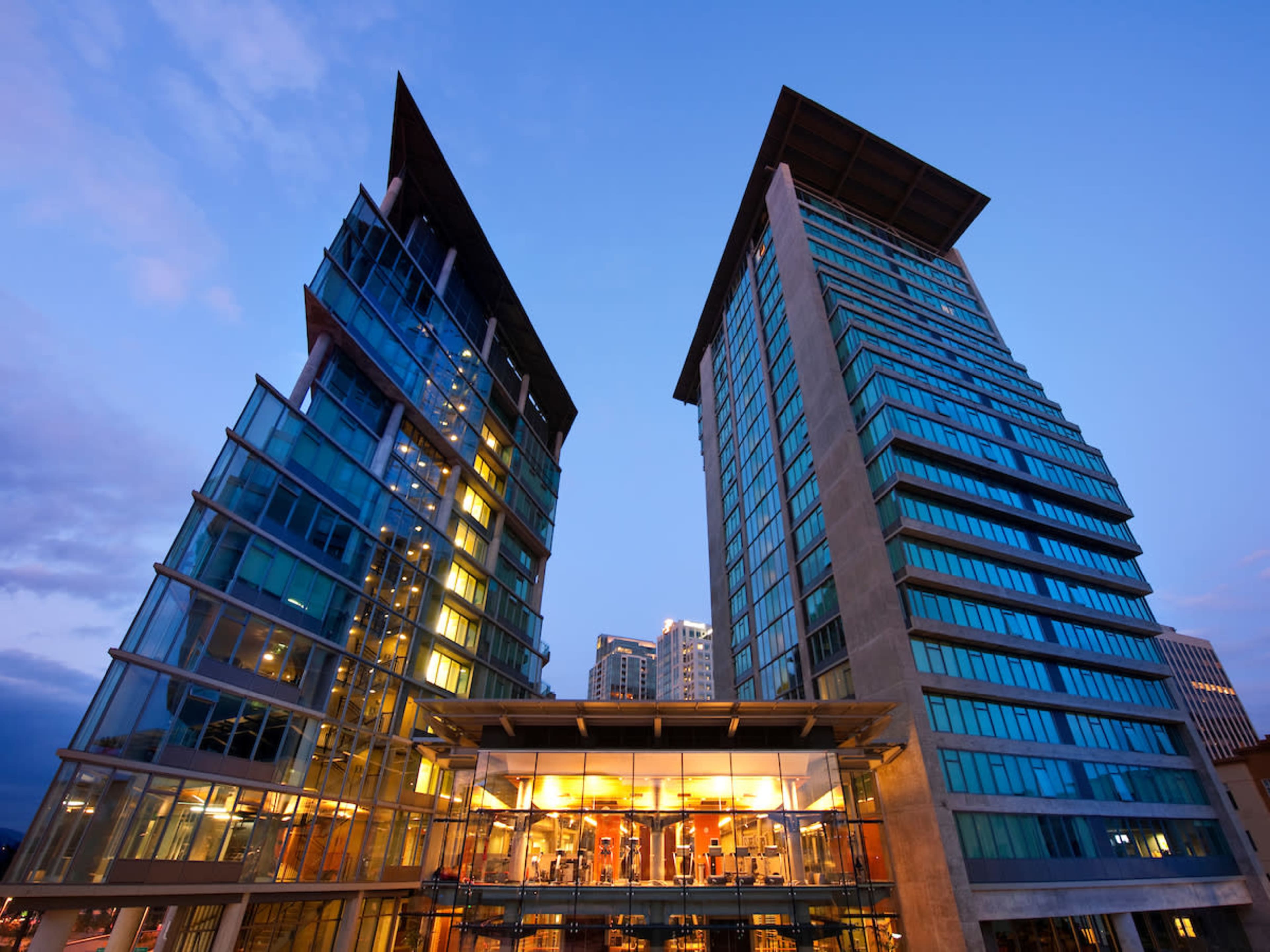 Two modern high-rise buildings with glass facades and a connected lower level illuminated at dusk.