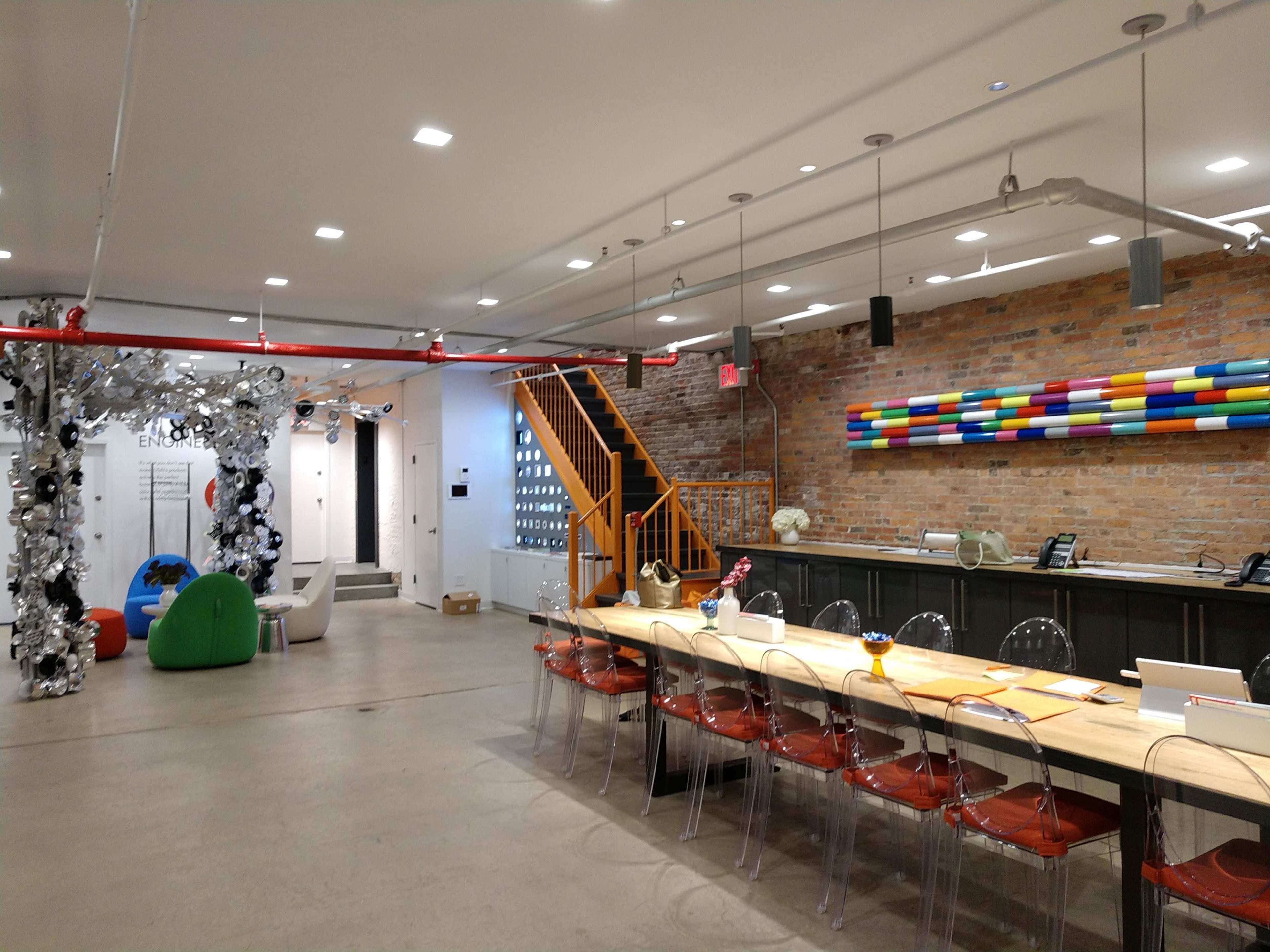 The image shows a modern, open office space with a long wooden table, clear chairs, a colorful wall art installation, and an entrance leading to another area.