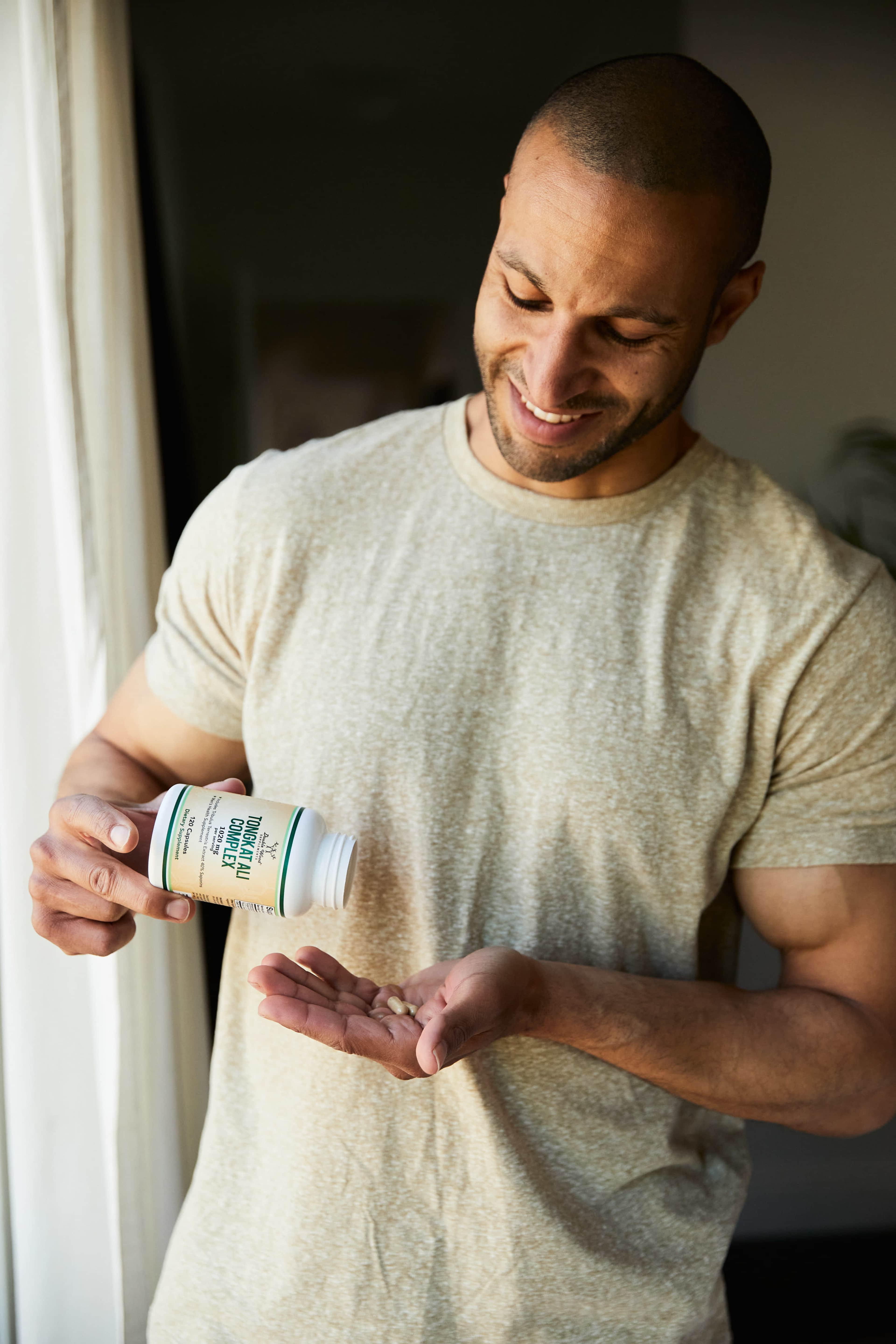 A man pours dietary supplements from a bottle into his palm while standing by a window.